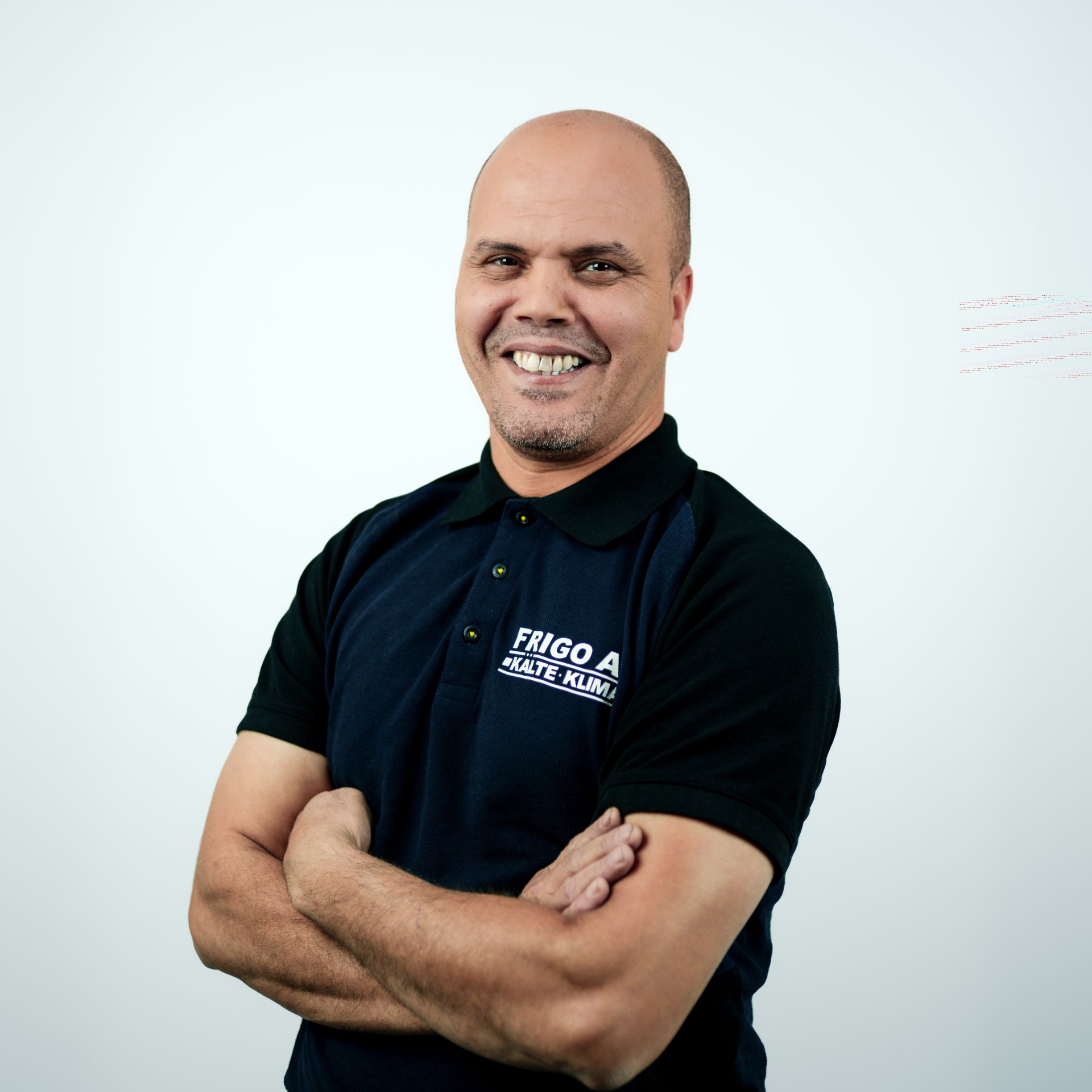 Mann mit verschränkten Armen, lächelnd; trägt ein schwarz-blaues Polohemd mit Logo und steht vor einem weißen Hintergrund.