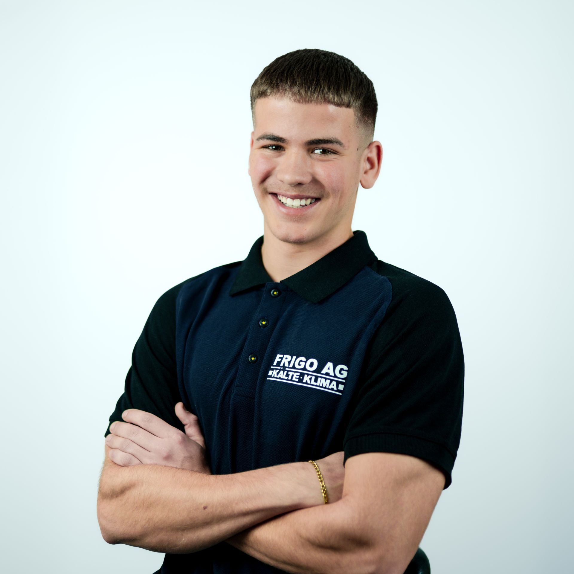 Ein lächelnder Mann mit verschränkten Armen trägt ein schwarz-blaues Poloshirt mit dem Logo „Frigo AG“. Weißer Hintergrund.