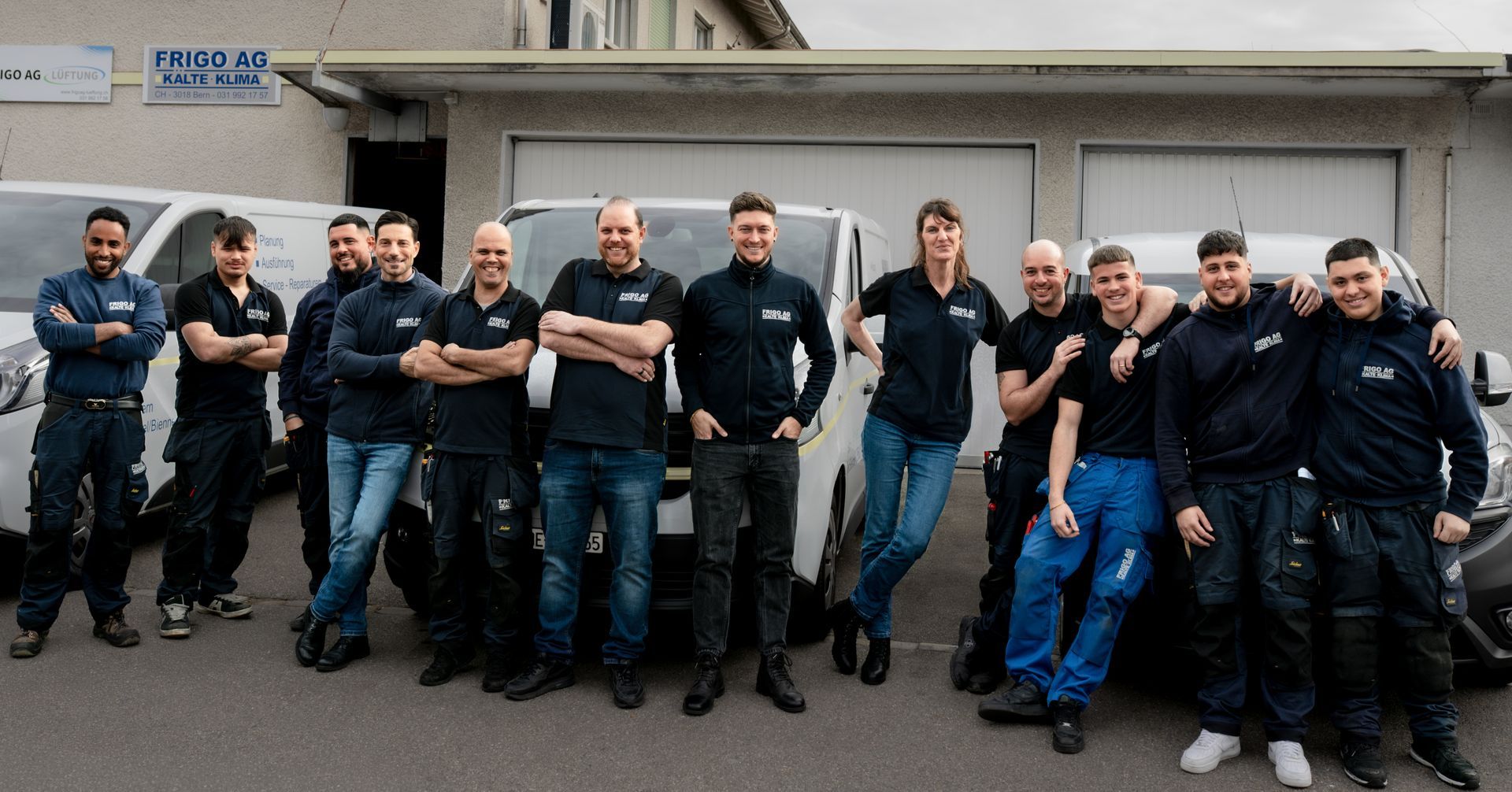 Eine Gruppe von Menschen posiert vor Lieferwagen vor einem Gebäude. Alle tragen Arbeitskleidung und lächeln.