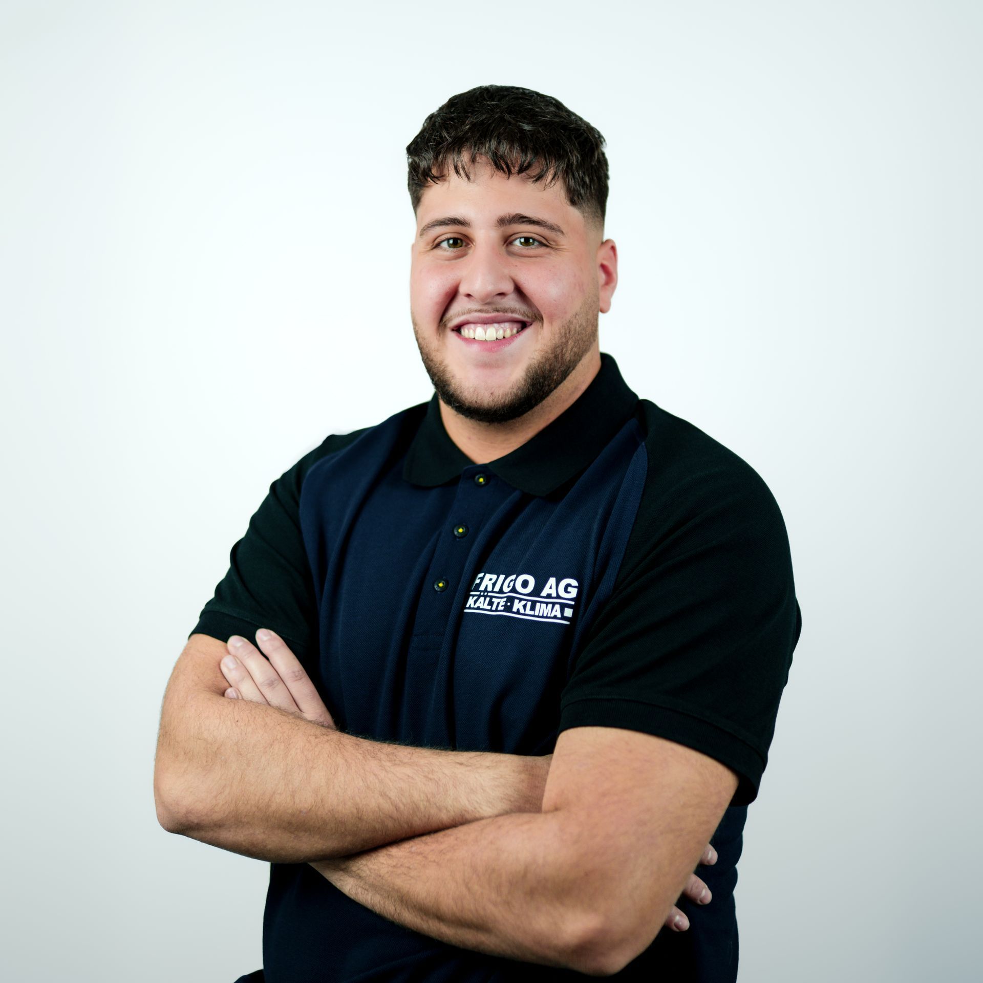 Mann mit verschränkten Armen, der ein dunkelblaues Poloshirt mit Logo trägt, lächelt vor einem weißen Hintergrund.