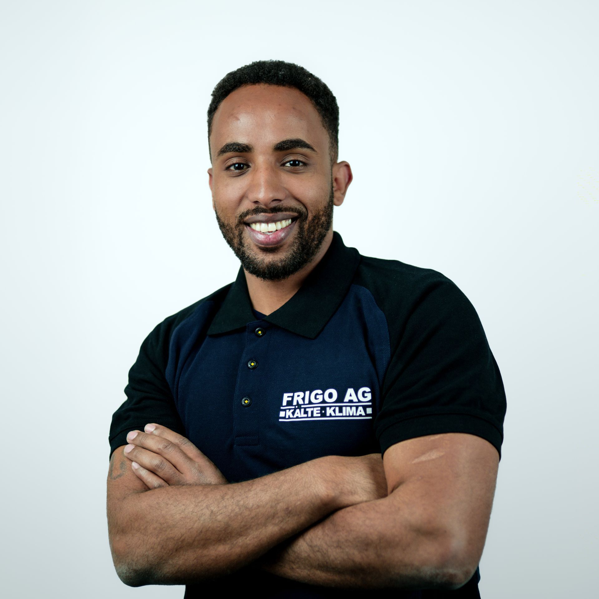 Ein lächelnder Mann mit verschränkten Armen, der ein marineblau-schwarzes Poloshirt mit dem Logo „FRIGO AG“ trägt.