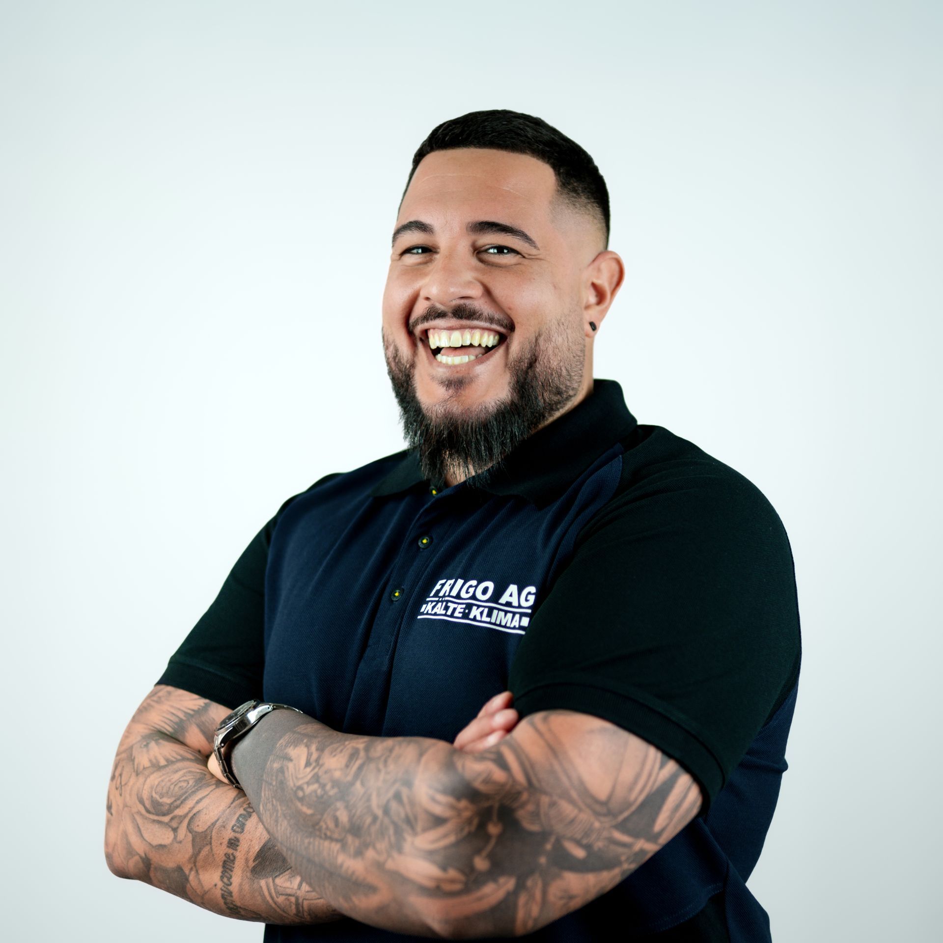 Ein Mann mit Bart und Tätowierungen lächelt mit verschränkten Armen und trägt ein dunkelblaues Poloshirt vor weißem Hintergrund.