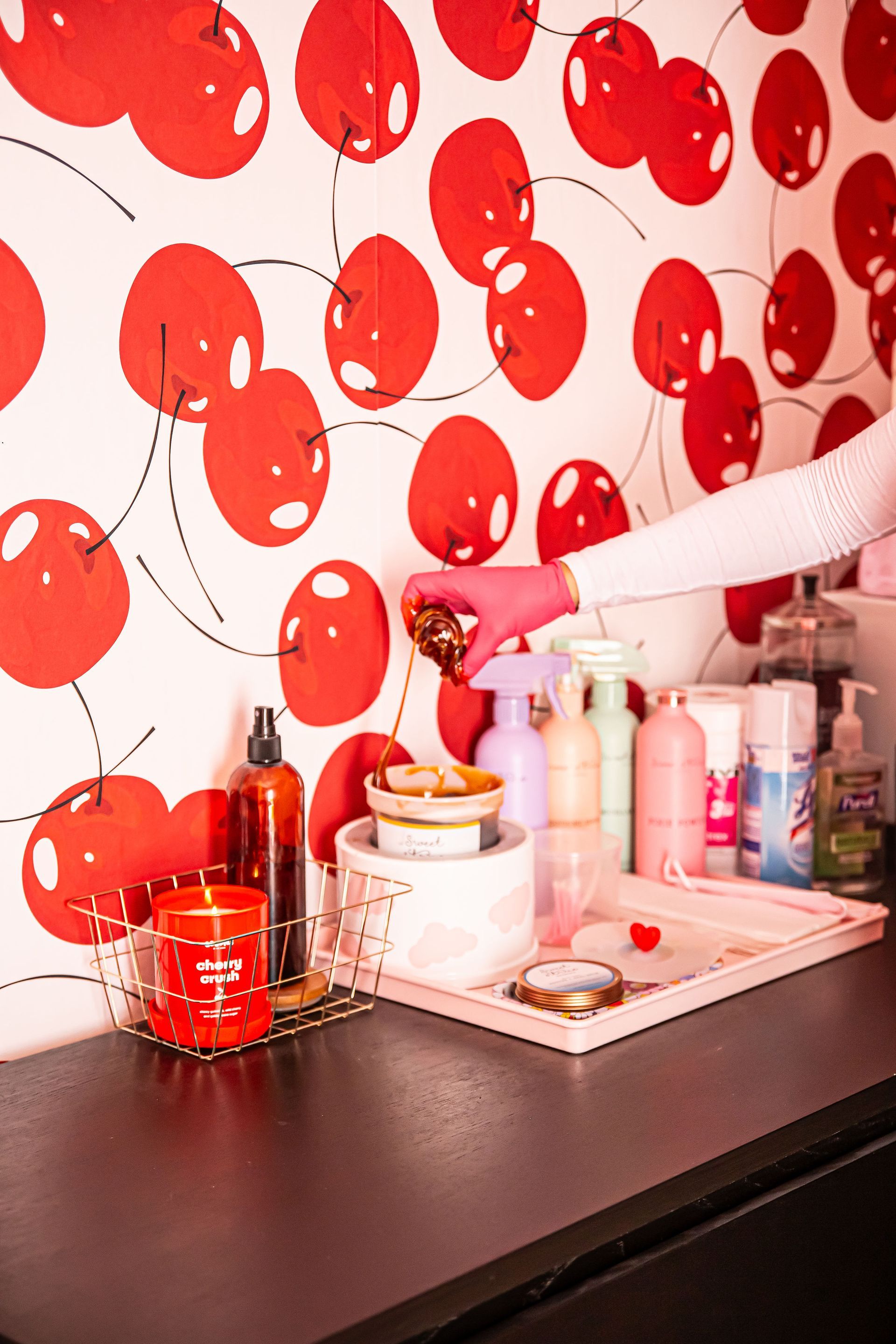 Person in pink gloves holding small item over beauty products, against cherry wallpaper background.