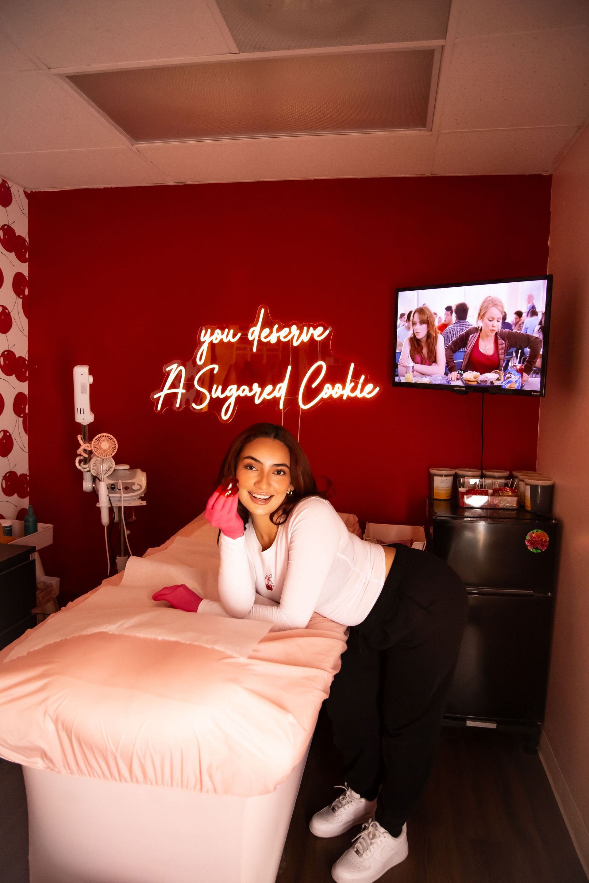 Woman in white top, black pants, leans on a pink-covered massage table in a pink room with a lit sign.