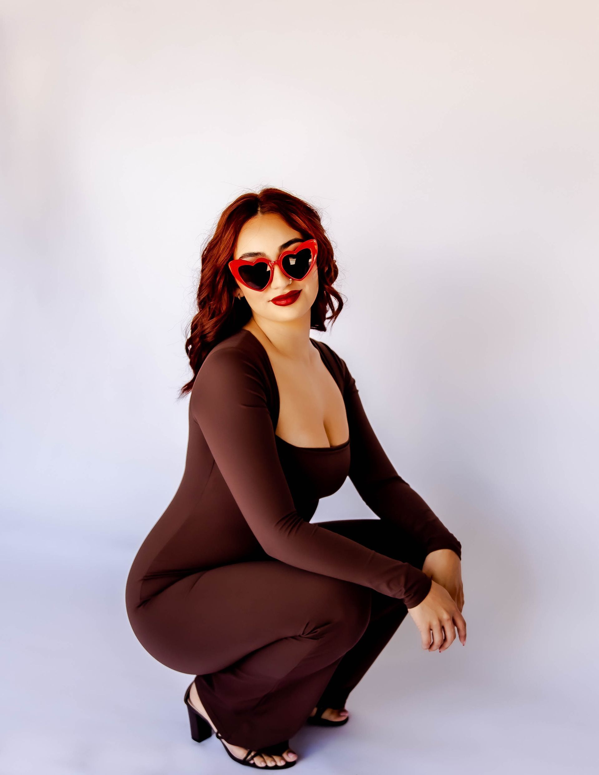 Woman in brown jumpsuit, red heart-shaped sunglasses, and high heels, squats against a white backdrop.