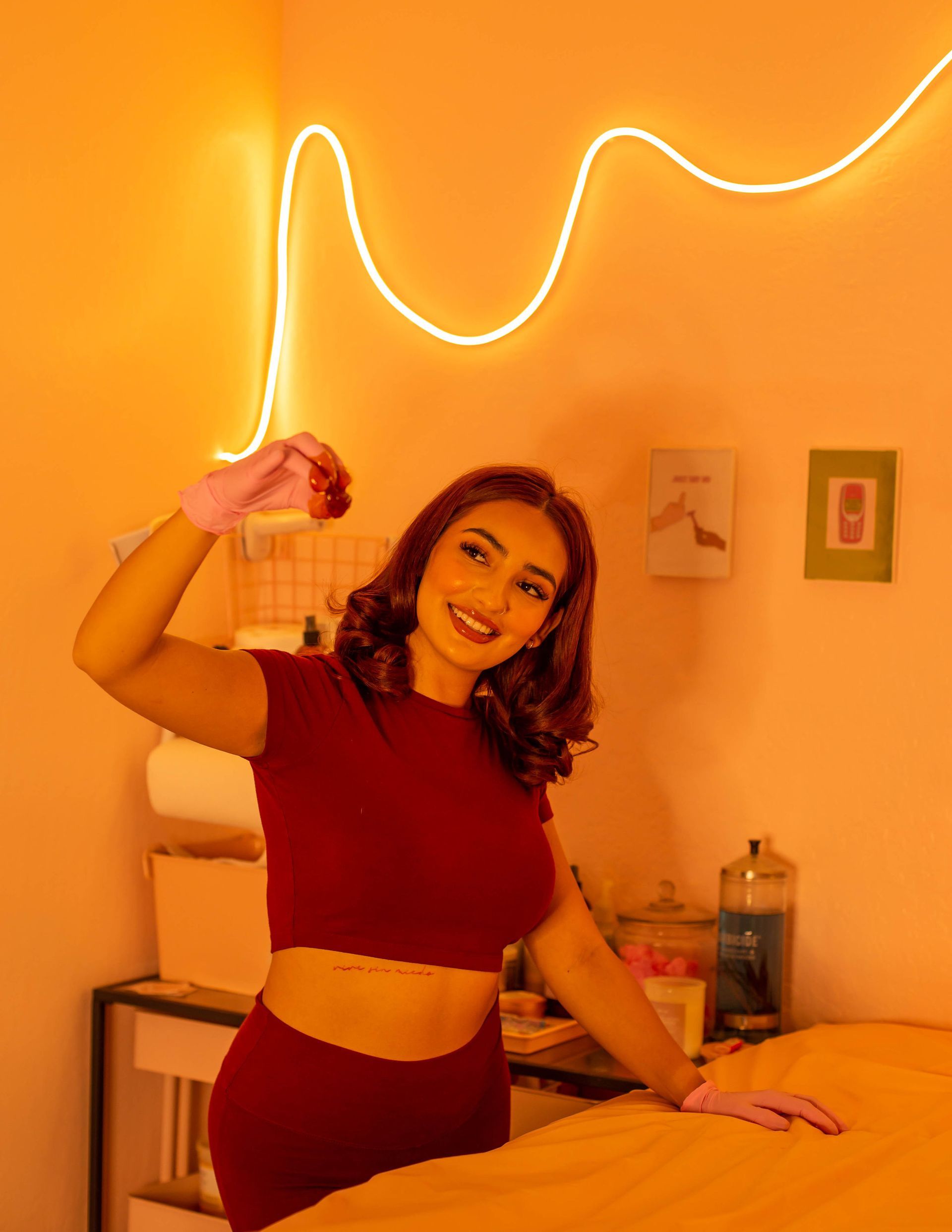 Woman in red, holding item, smiling near a massage table with neon sign, in a room with light orange tones.
