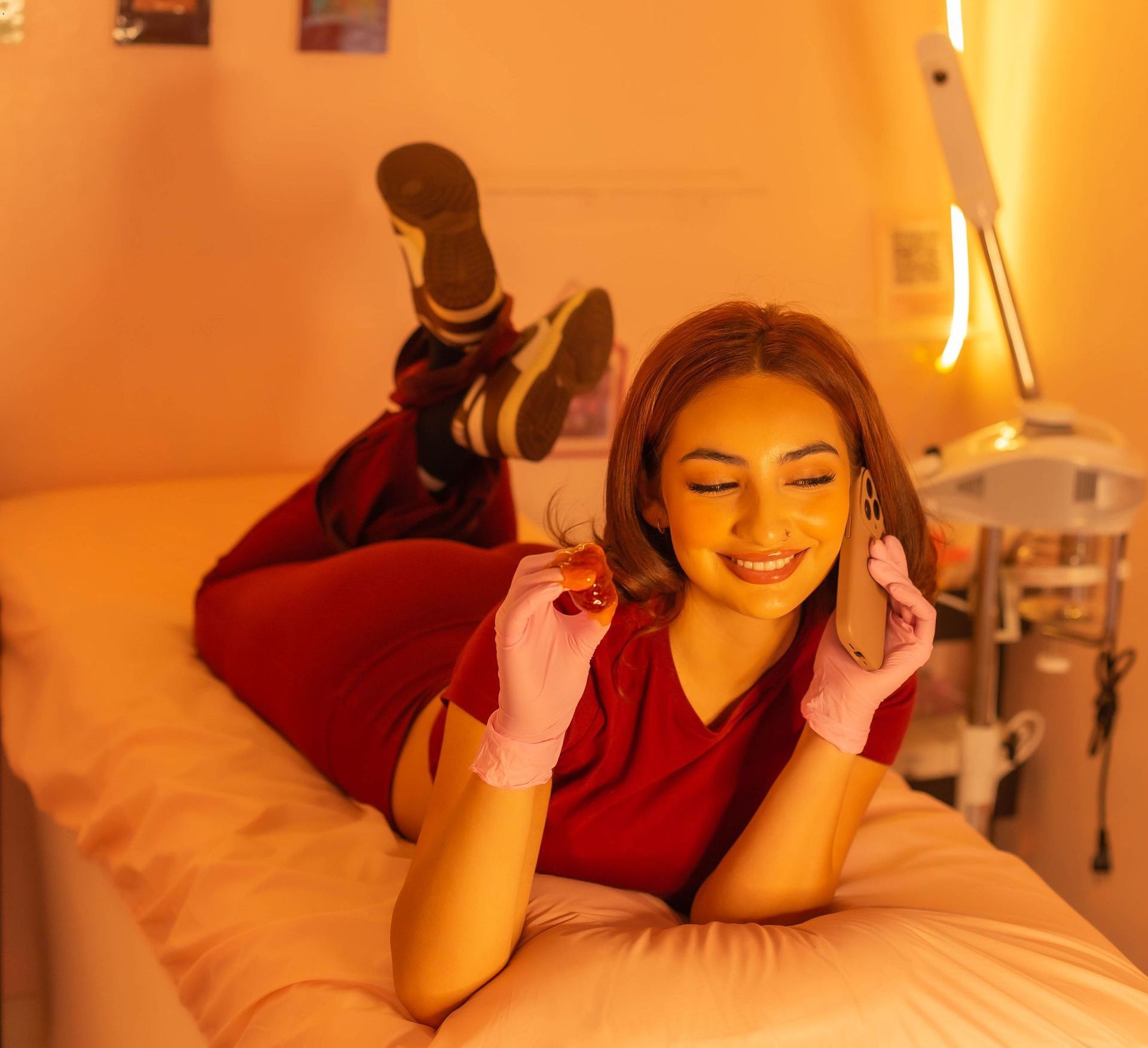 Woman reclines on a massage table, holding a phone and lash extensions. Pink-gloved hands, red outfit, warm-lit room.
