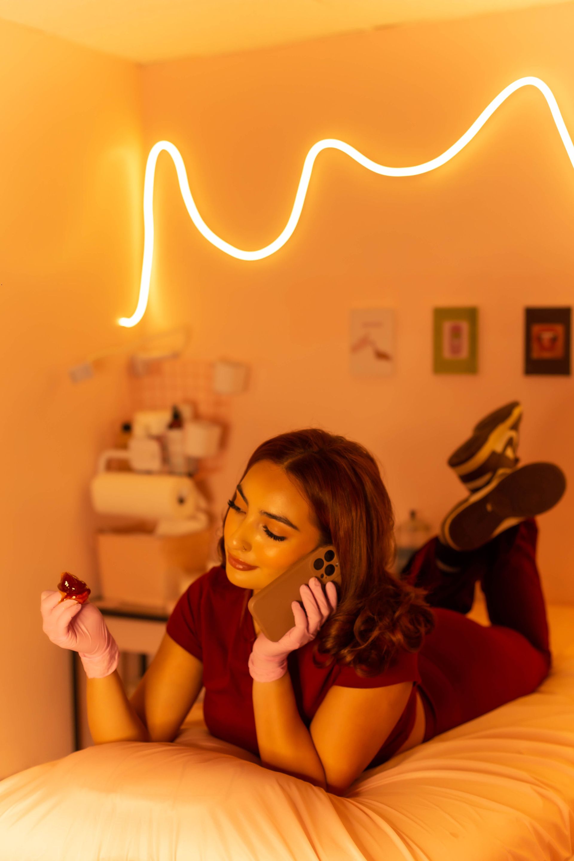Woman lying on a massage table, on the phone, holding a pen. Pink-lit room with neon sign.