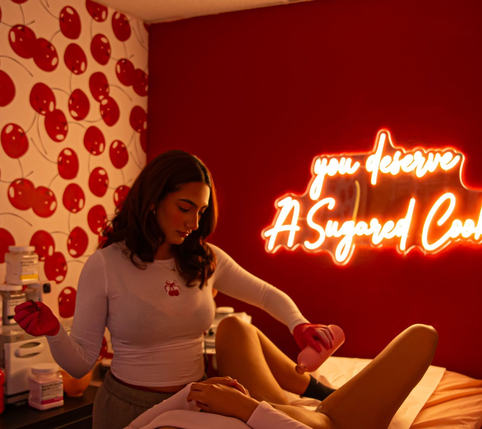 Woman applying wax to someone's legs in a pink-themed room with a neon sign that says, 