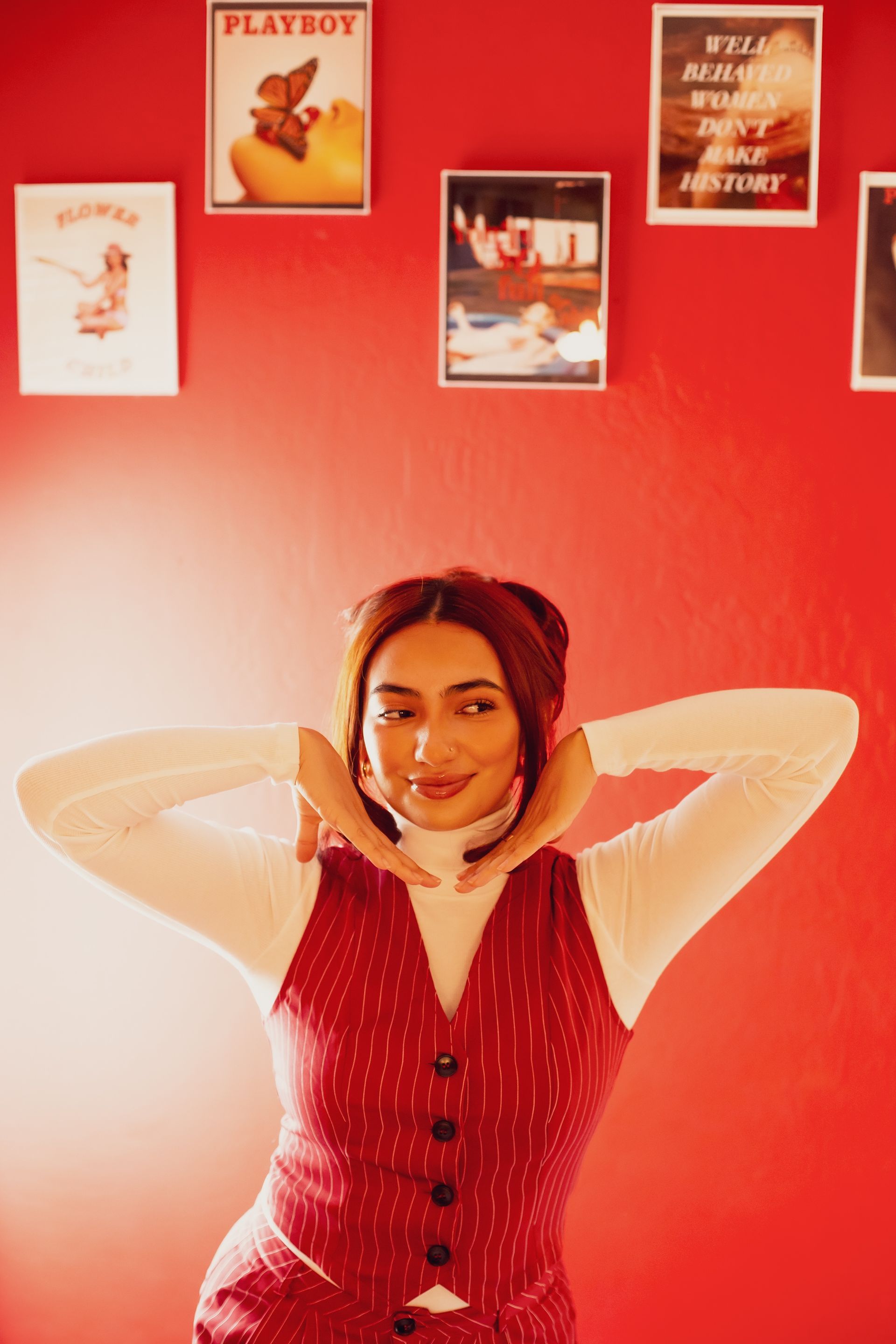 Woman in a red vest and white turtleneck poses with hands framing her face against a red wall with framed pictures.