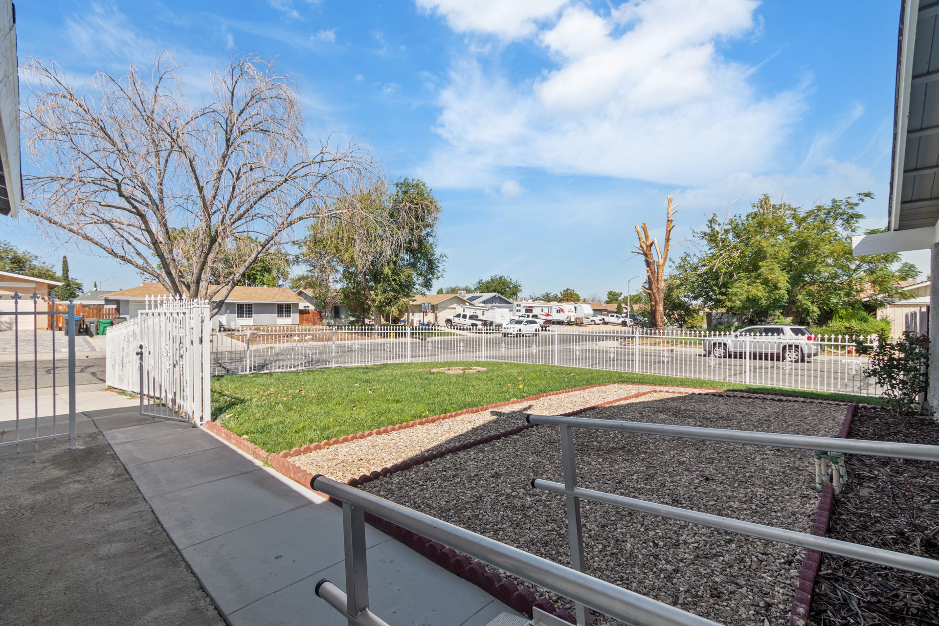 View from doorway of a yard with grass, bushes, a white fence and street with cars under a blue sky.