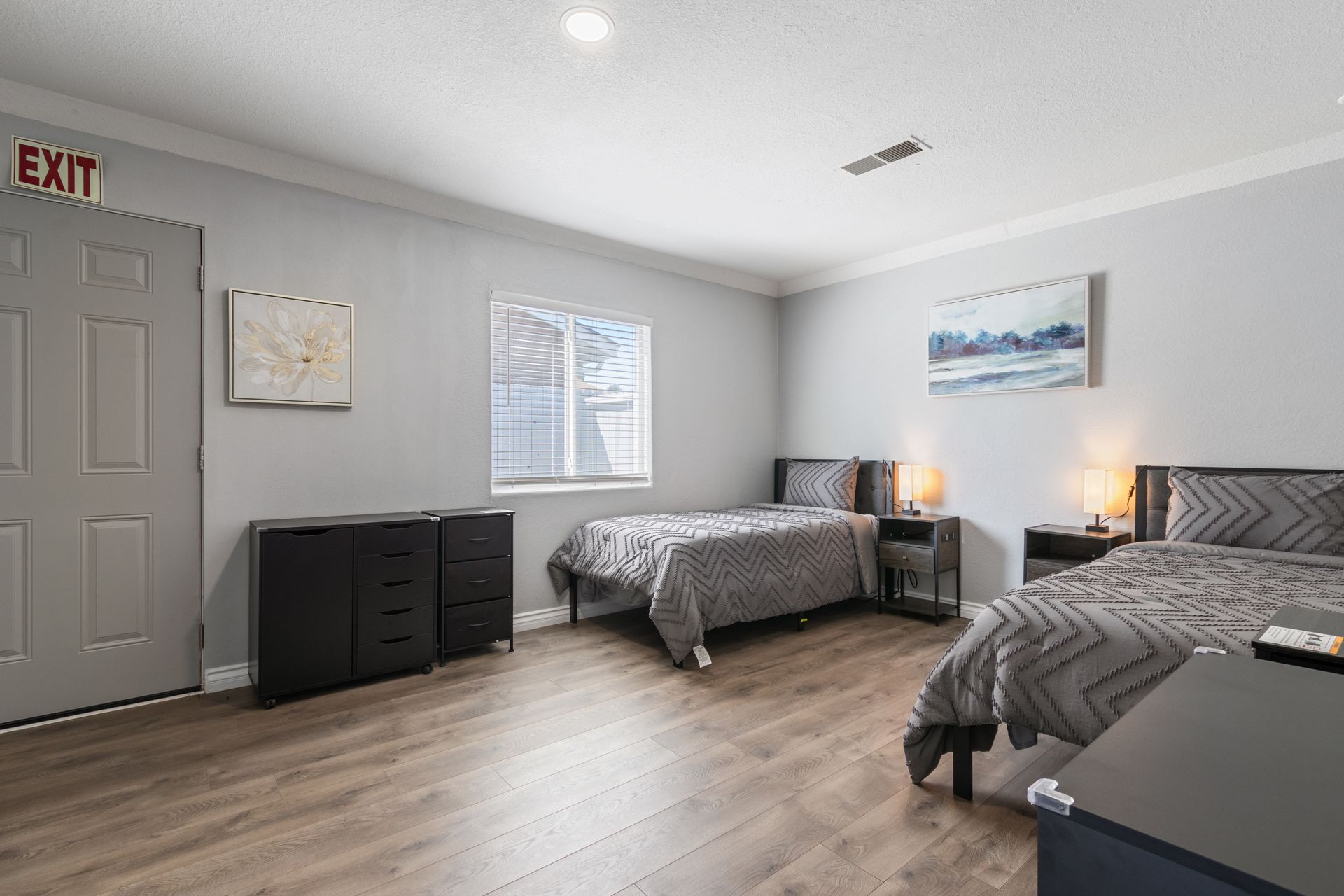 A bedroom with two beds, dressers, and an exit door. Gray walls and wood-like flooring.