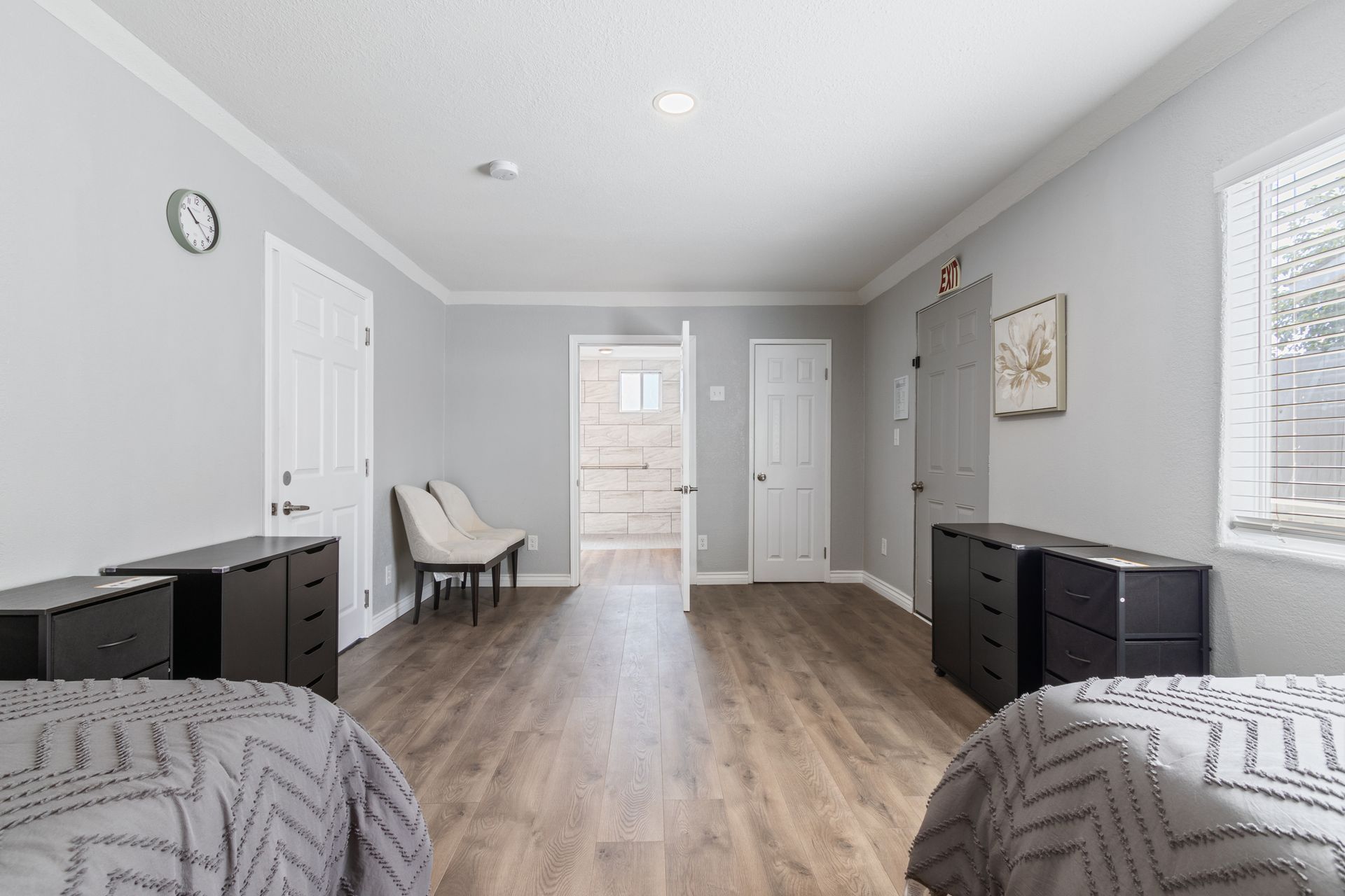 An interior room with two beds, dark dressers, two chairs, and a hallway. Gray walls, wood floor.