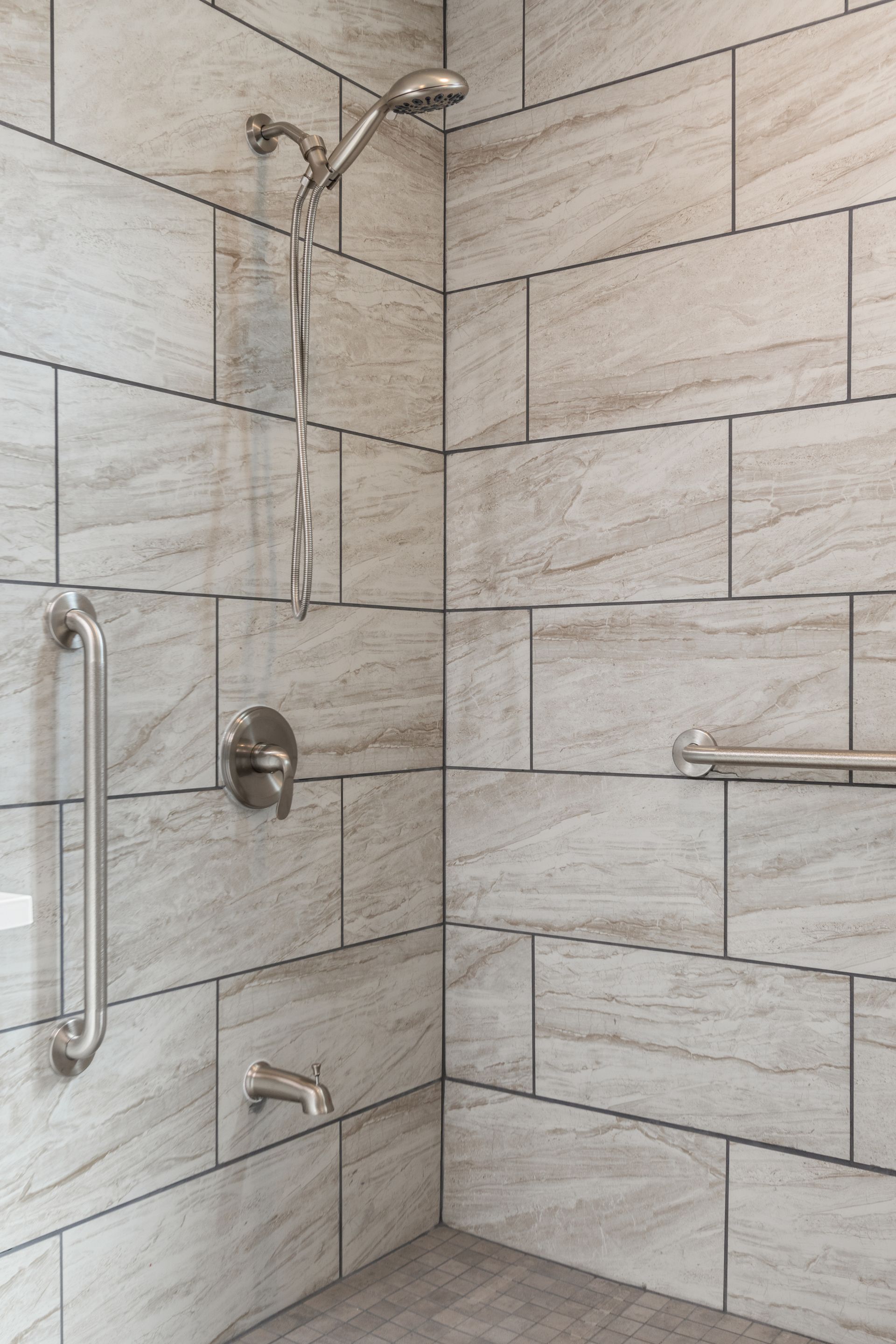 Tiled shower with grab bars.  Silver fixtures and stone-look tiles.
