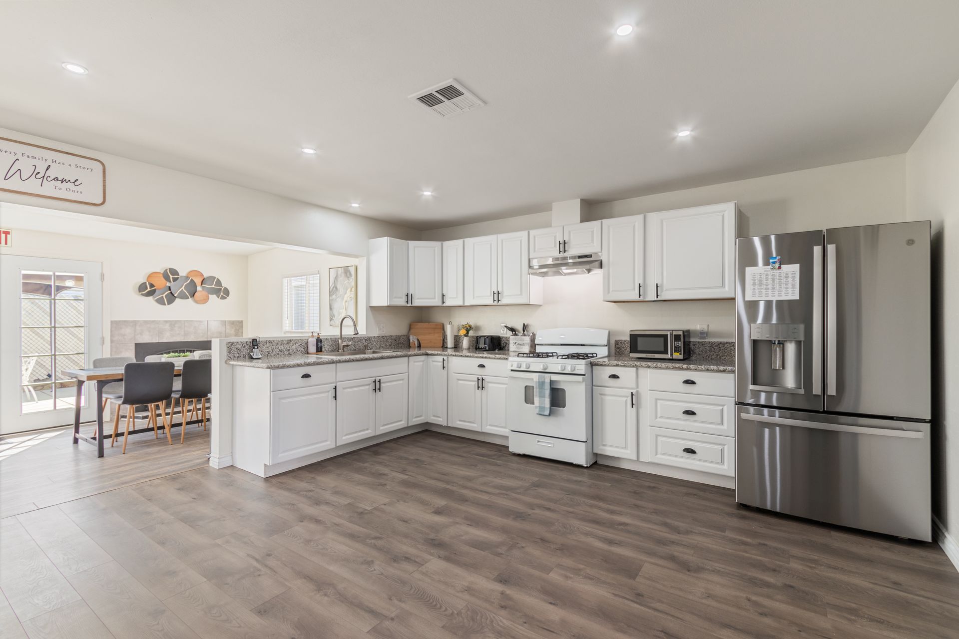 Spacious white kitchen with stainless steel refrigerator, gas stove, and light wood flooring. Dining area visible on left.