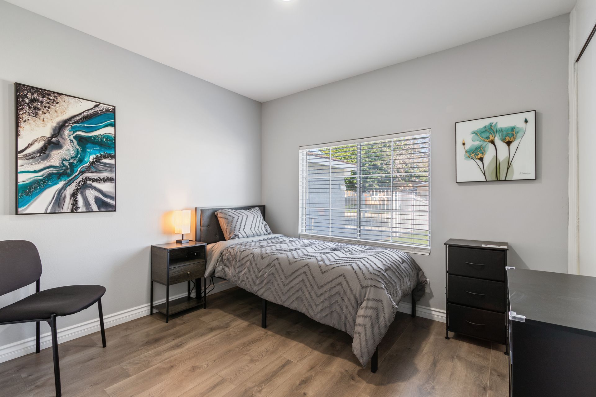 Bedroom with twin bed, nightstand, chair, dresser, artwork, and a window with blinds.