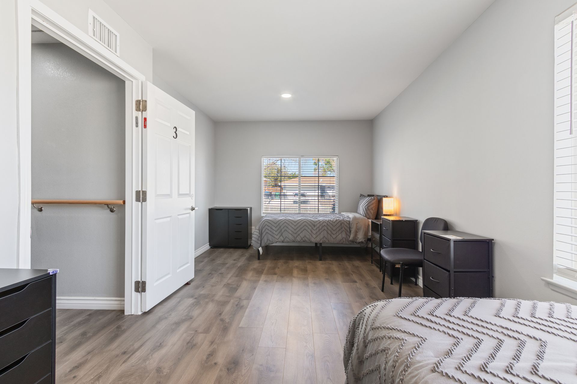 A bedroom with two beds, a closet, and a window. Gray walls, wooden floors, and minimalist furniture.