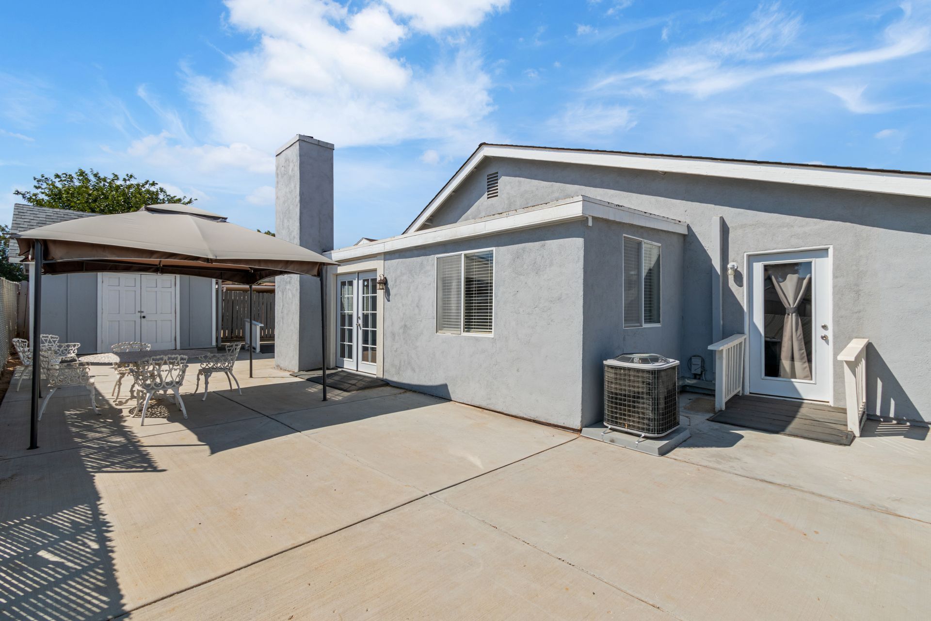 A gray house with a concrete patio, canopy, and storage shed on a sunny day.
