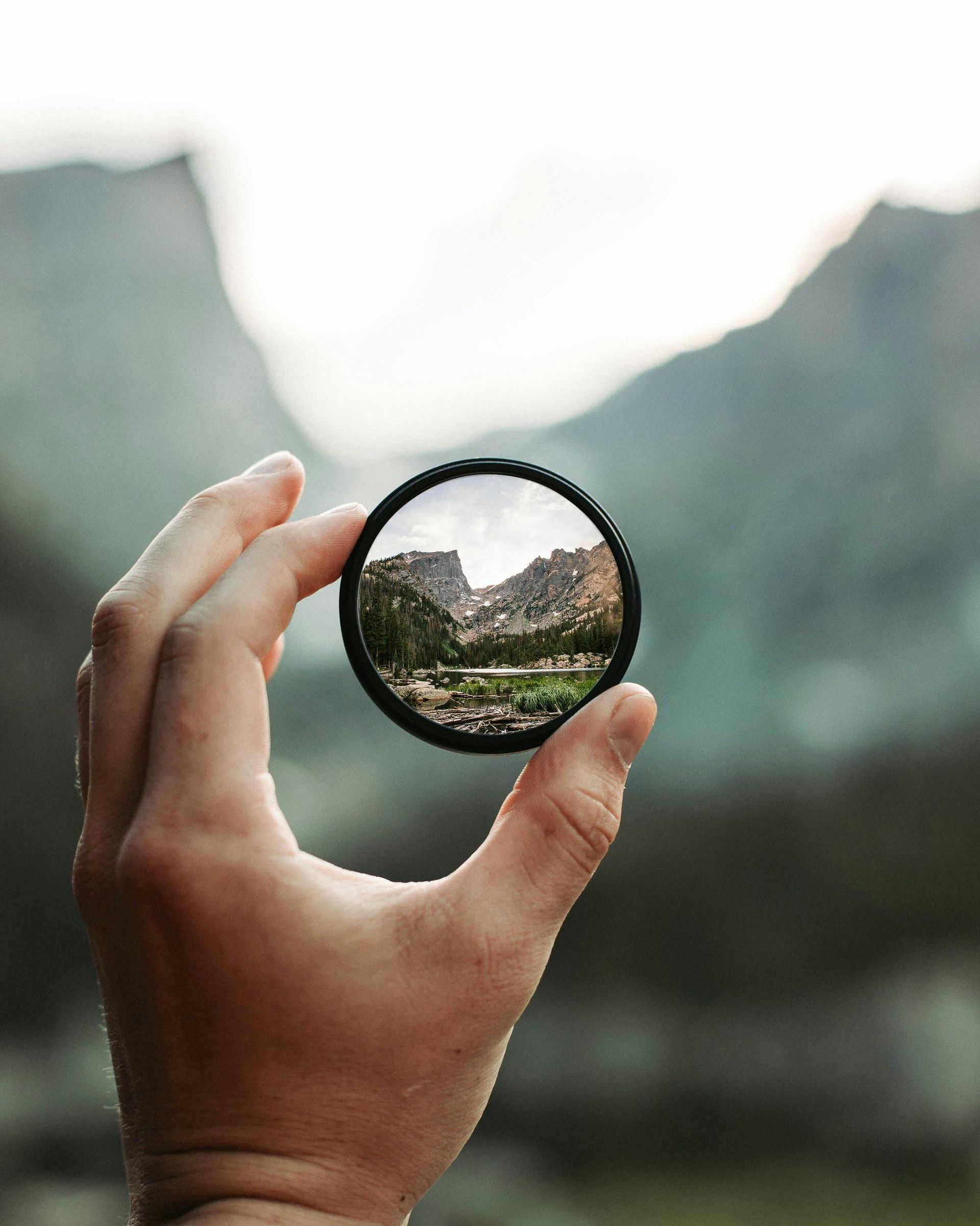 Hand holding a circular lens framing a mountain landscape.