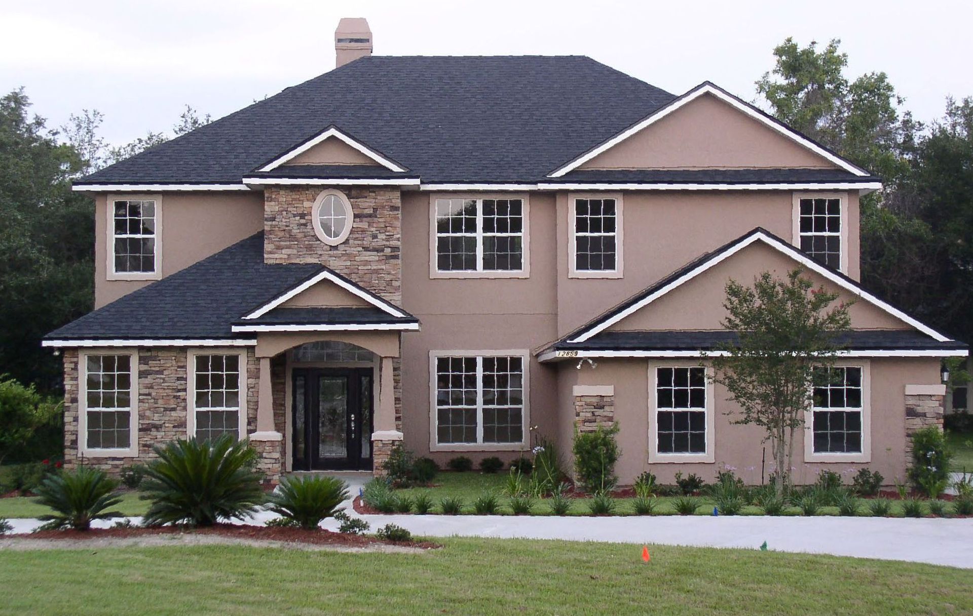 Two-story tan house with black roof, stone facade, and manicured lawn.
