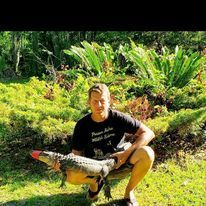 A man is squatting down in the grass holding a lizard.