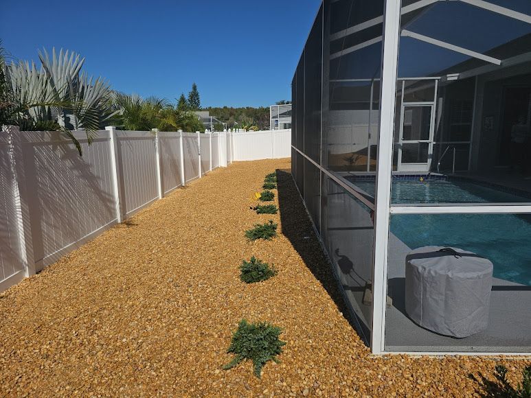 Narrow gravel side yard with small plants beside a screened patio and white fence under a blue sky