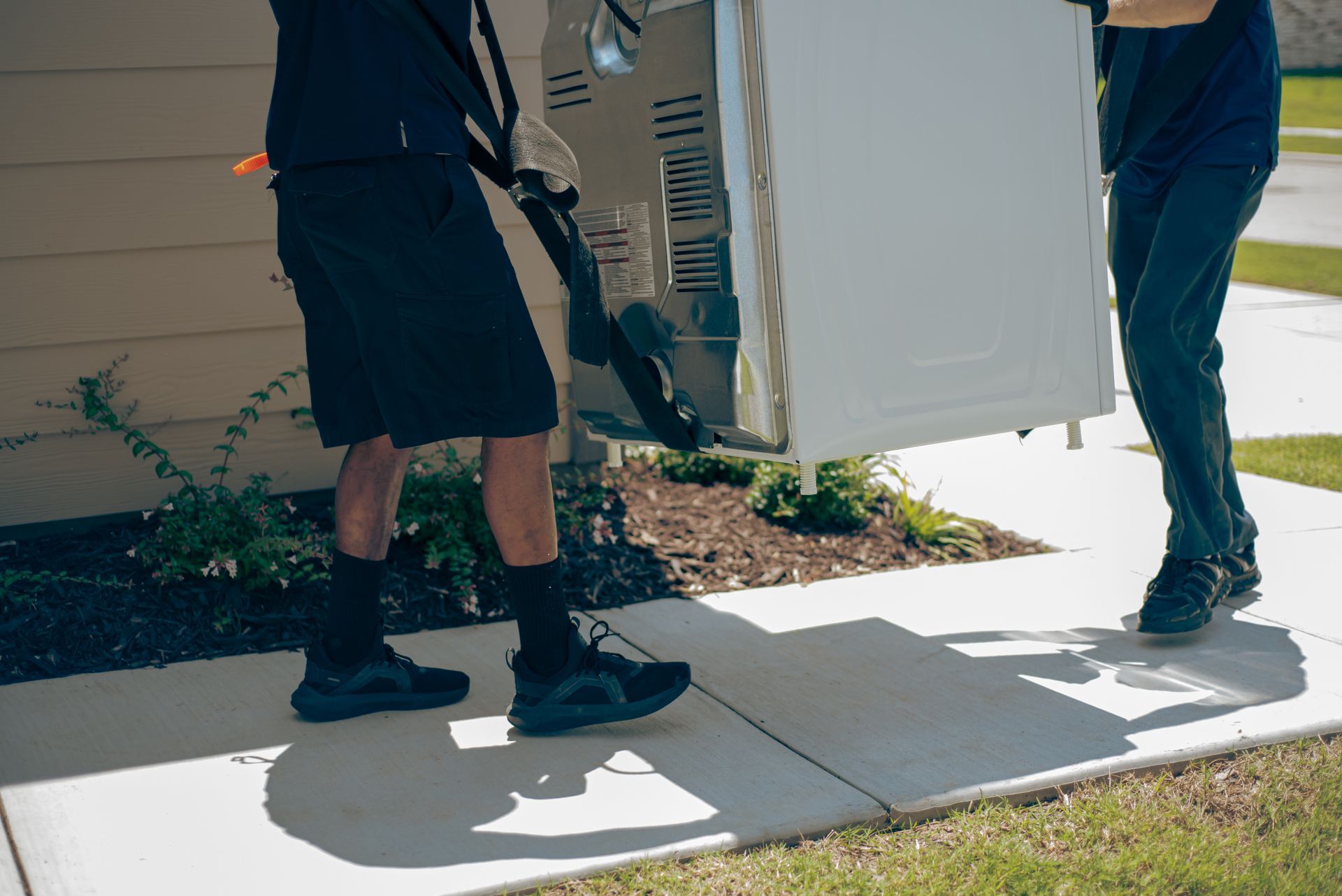 Two workers carrying a white appliance across a sidewalk into a house