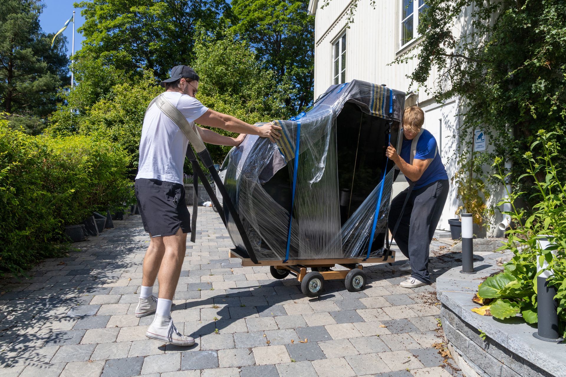 Two people moving a large wrapped item on a cart along a stone driveway beside a house.