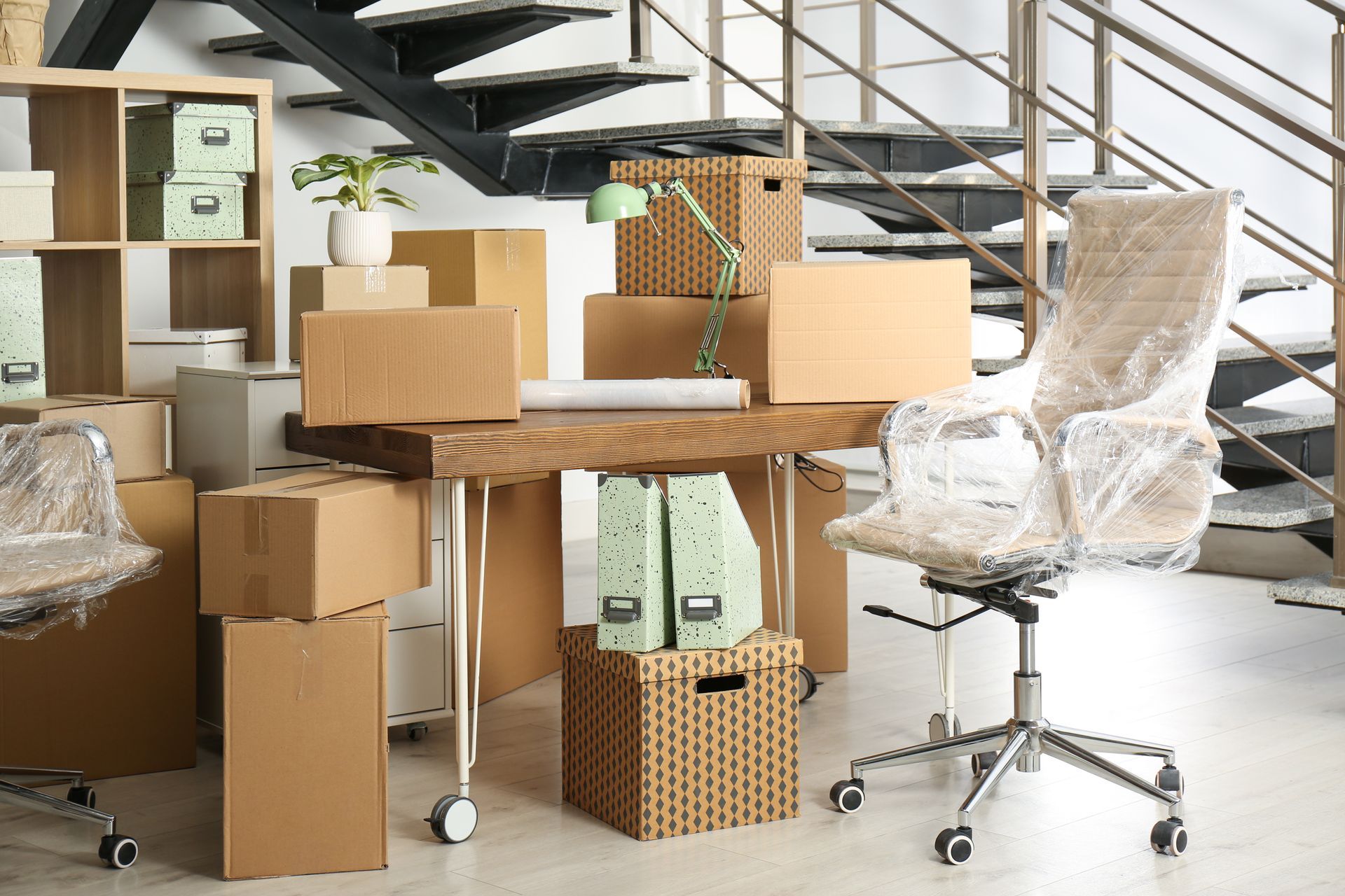 Moving boxes and wrapped furniture in an office, with a chair and staircase in the background.