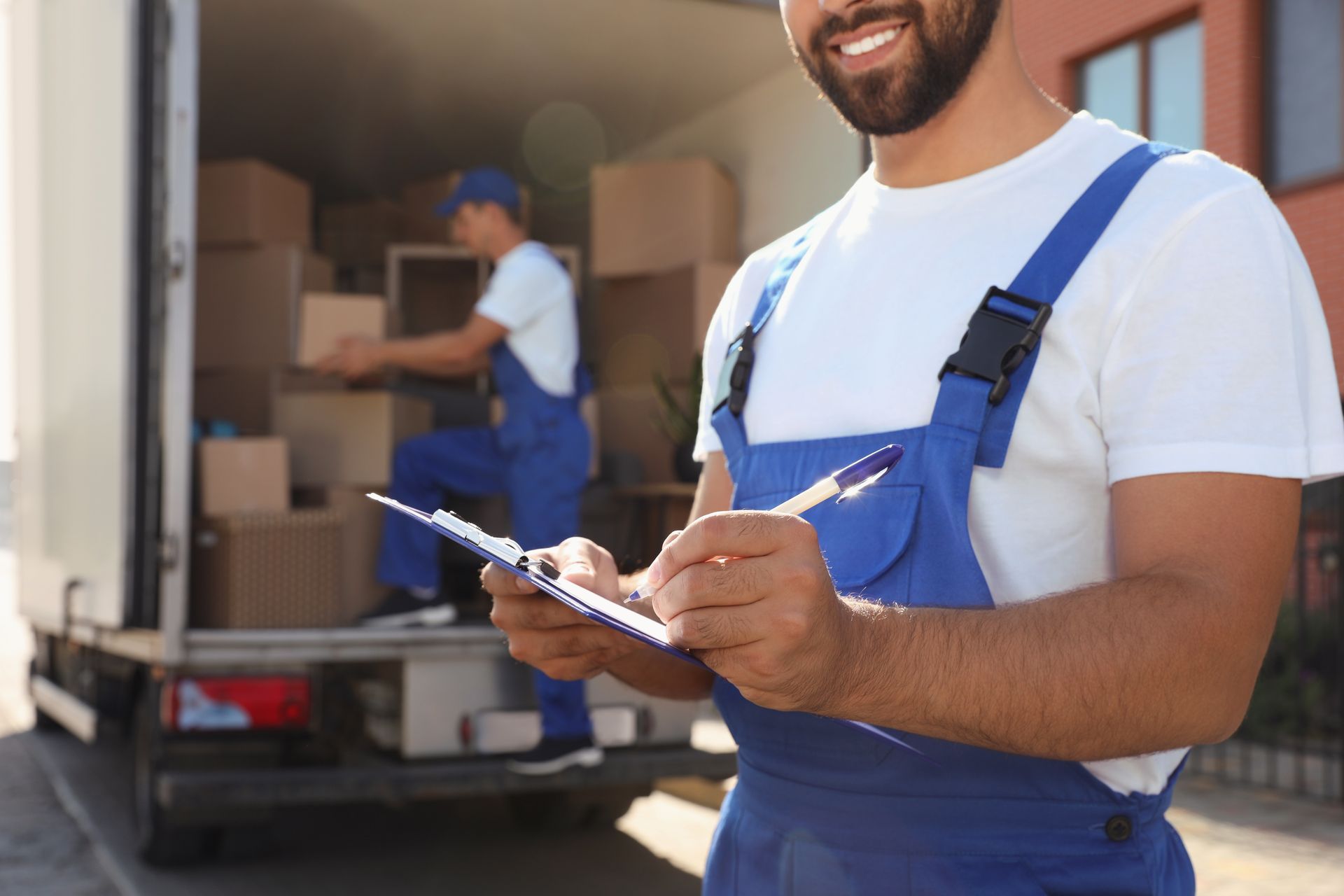 Delivery worker in blue overalls checking a clipboard beside an open moving truck