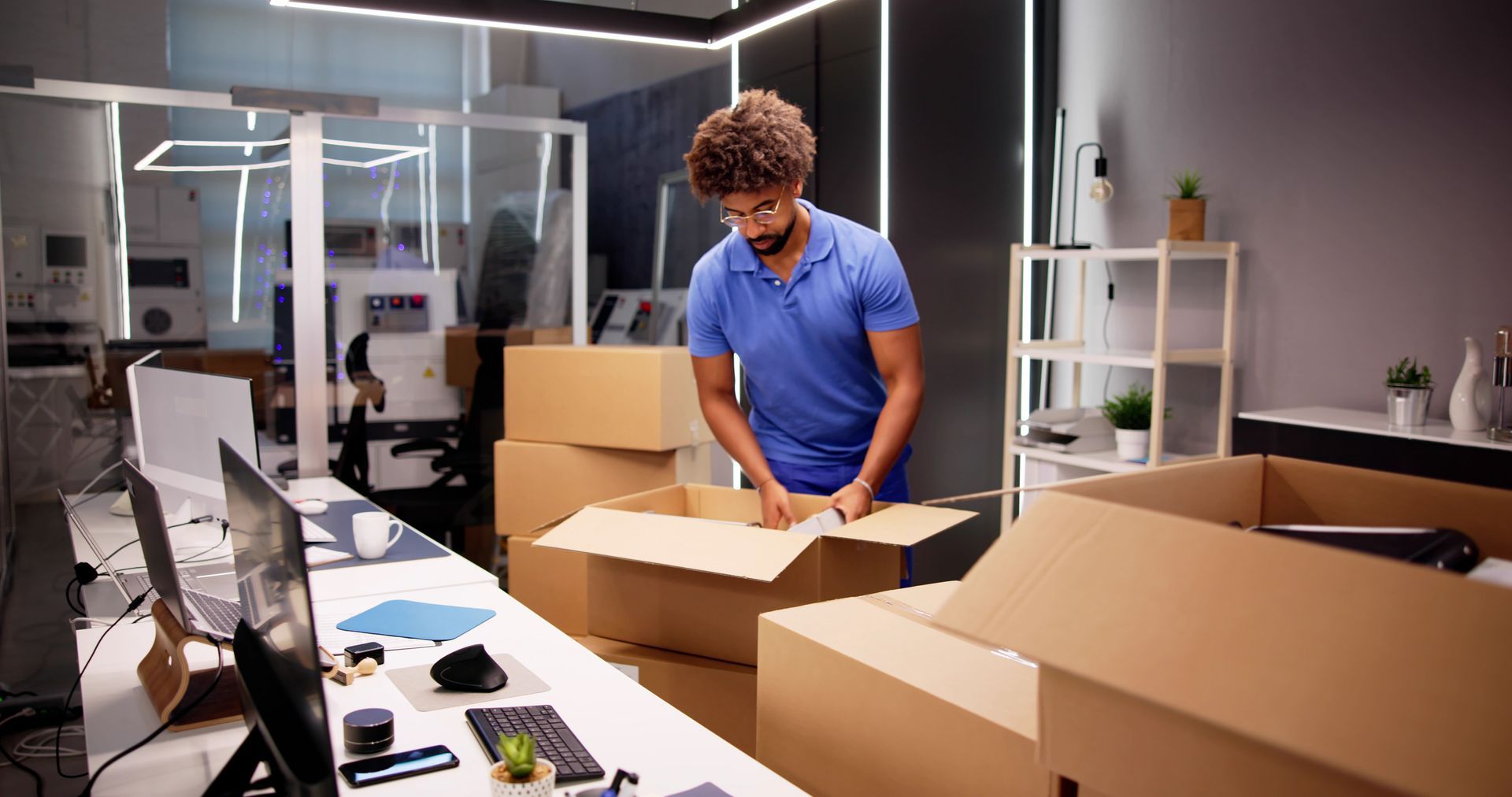 Person packing cardboard boxes in an office workspace with laptops and shelves in the background