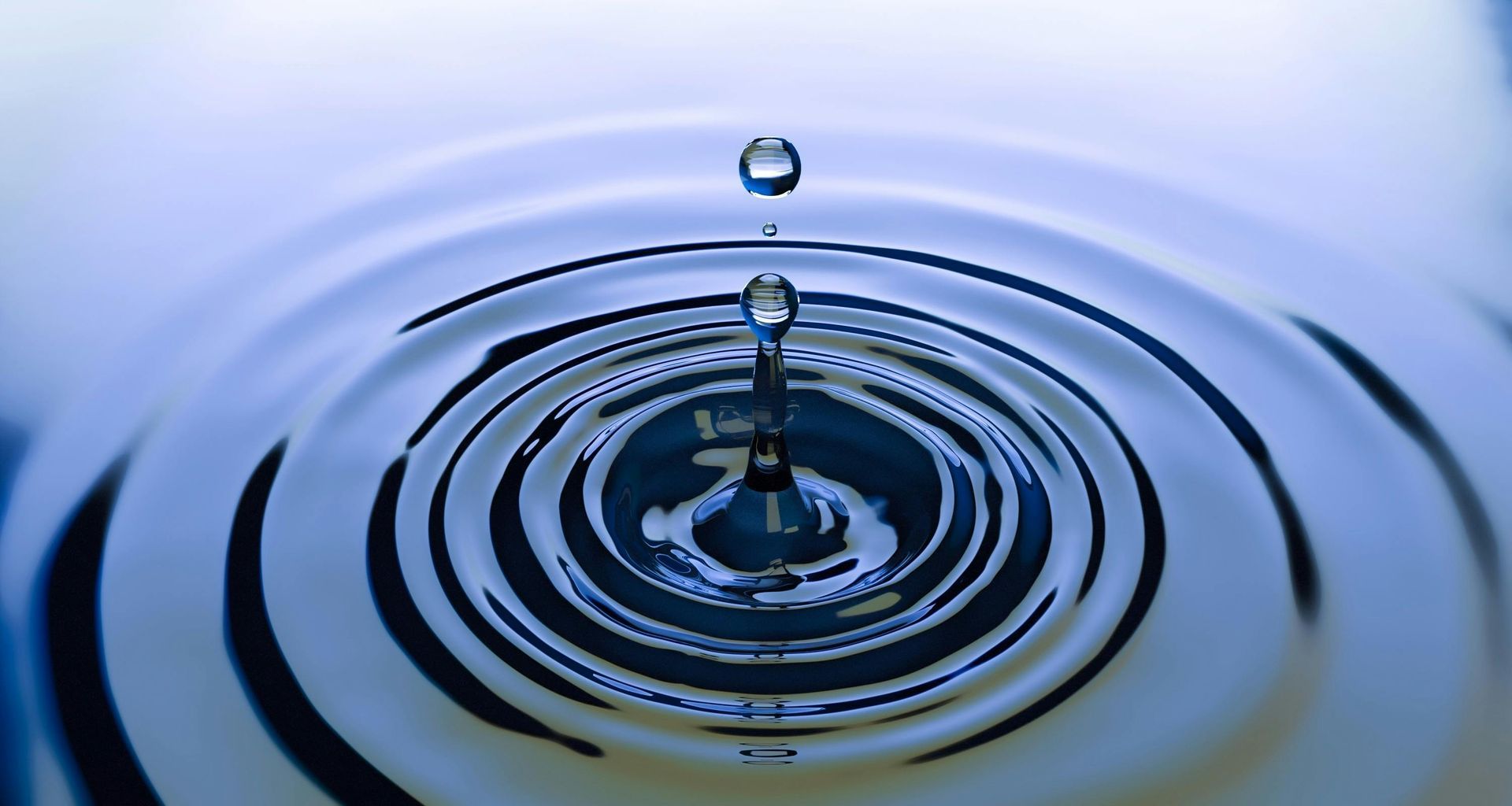 A water droplet suspended above concentric ripples in a blue-toned liquid.