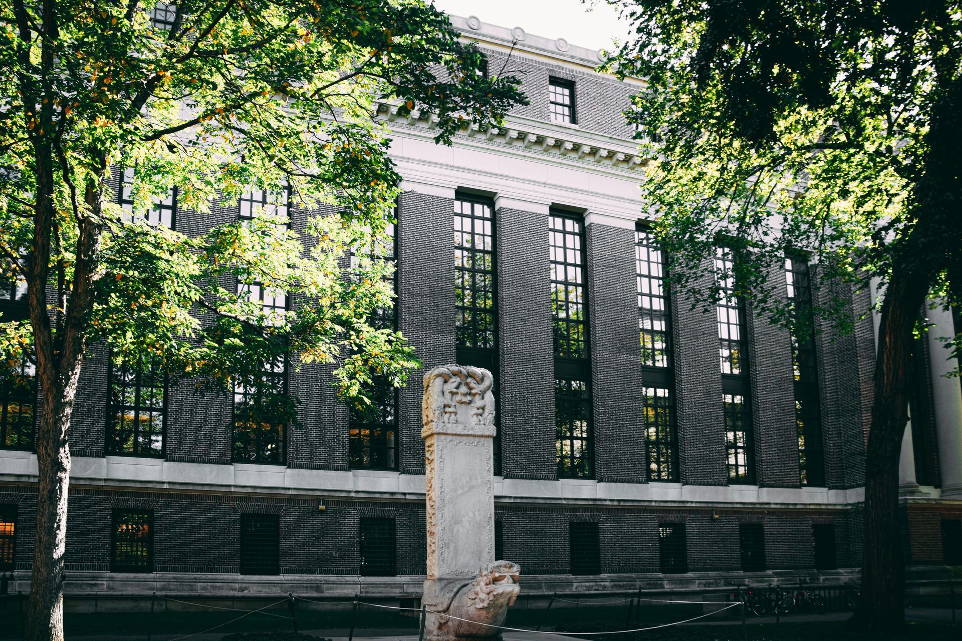 Brick building with tall windows framed by trees; a decorative stone column in the foreground.
