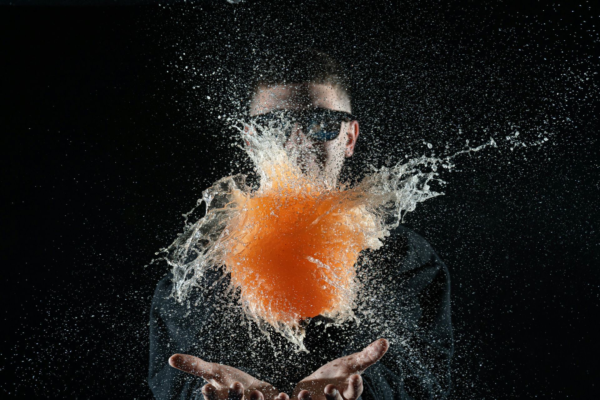 A person wearing sunglasses stands behind a bursting orange water balloon, creating a spray of droplets against black.
