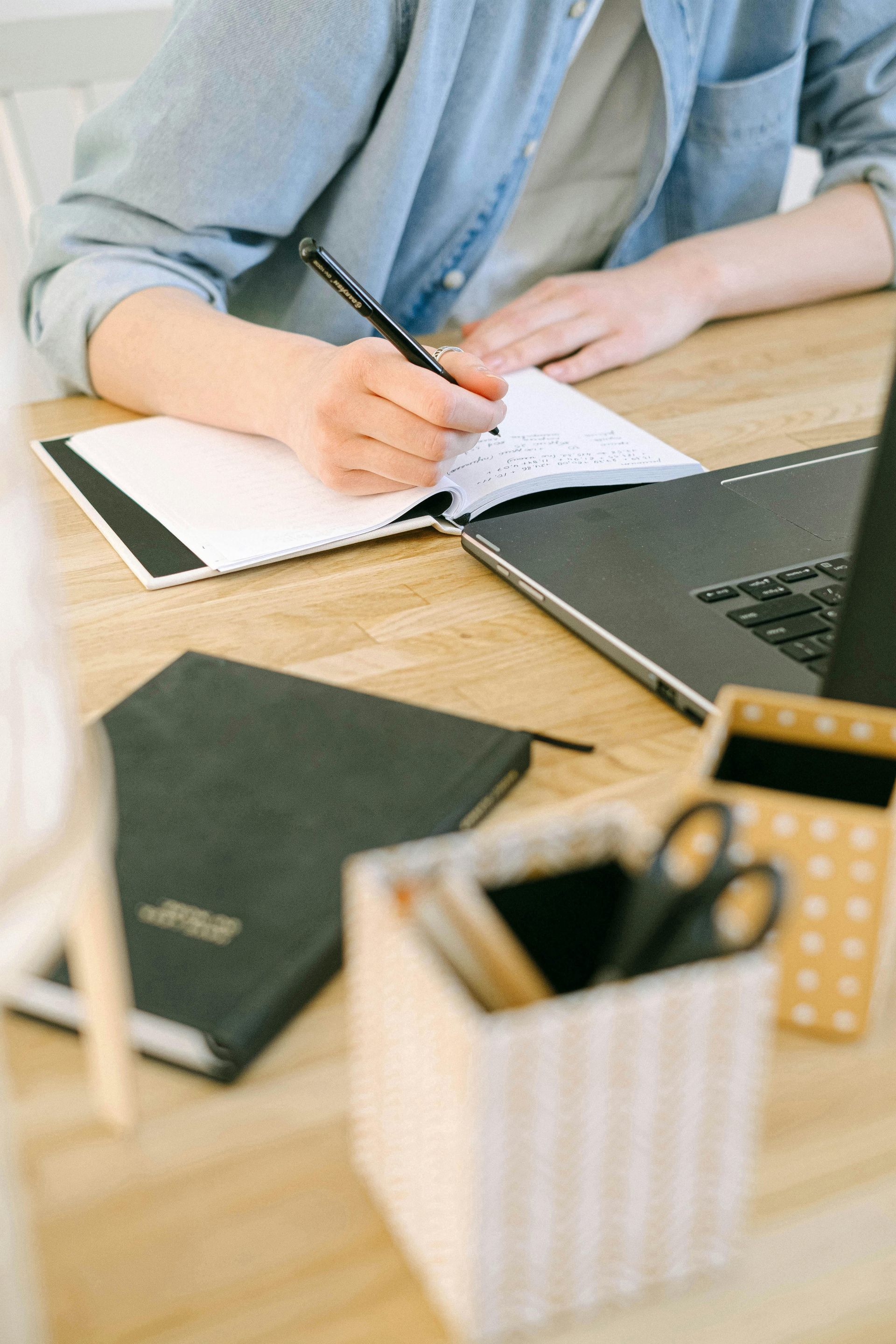 Person writing in a notebook at a wooden desk with a laptop, planner, and supplies.