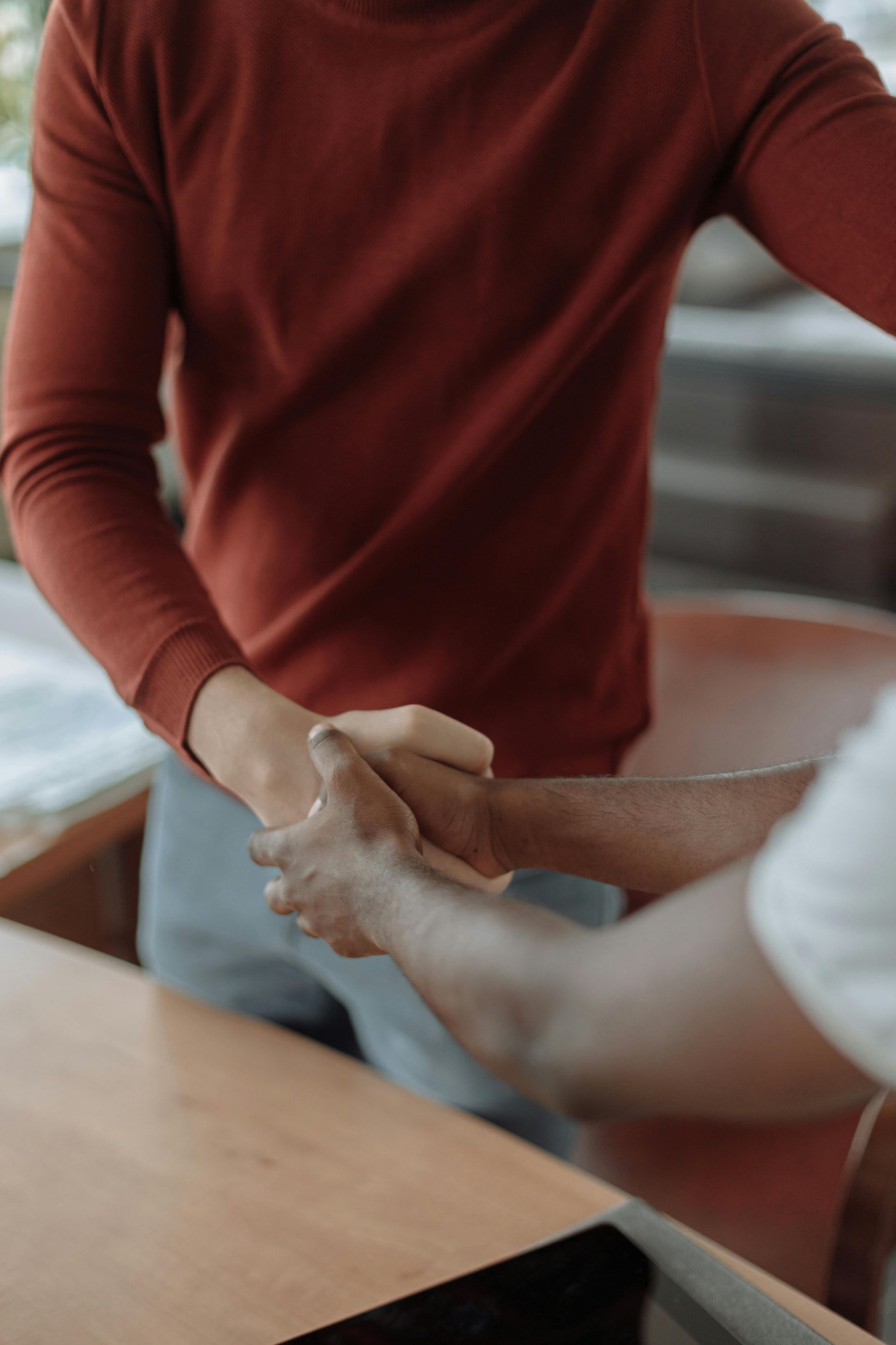 Two people shaking hands over a wooden surface; one in a red sweater, one in a white shirt.