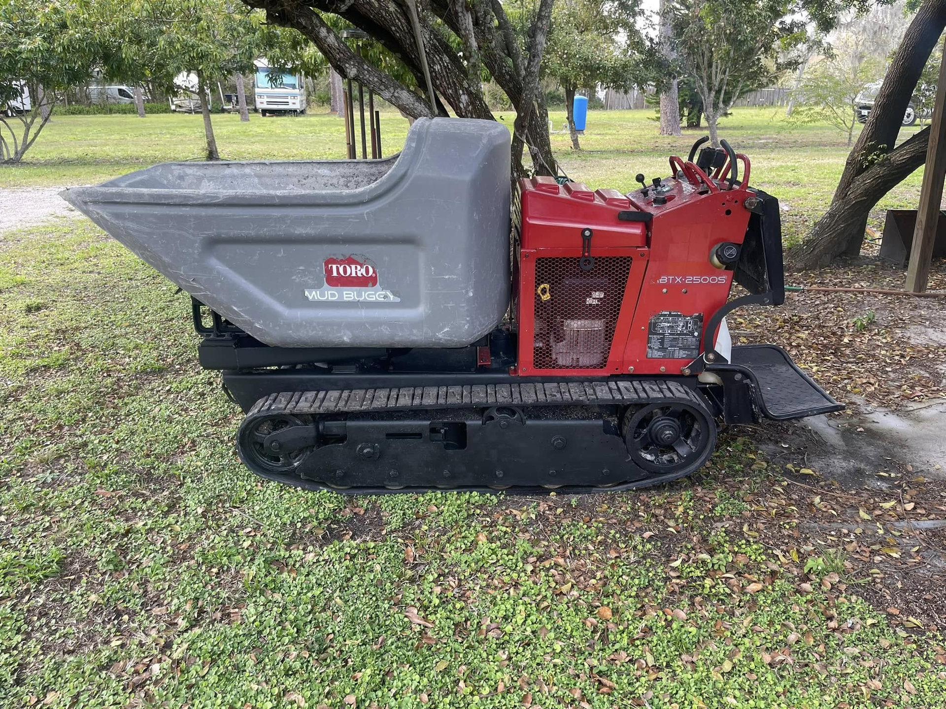 Toro tracked dumper on grass, red and gray, under trees near a camping area.