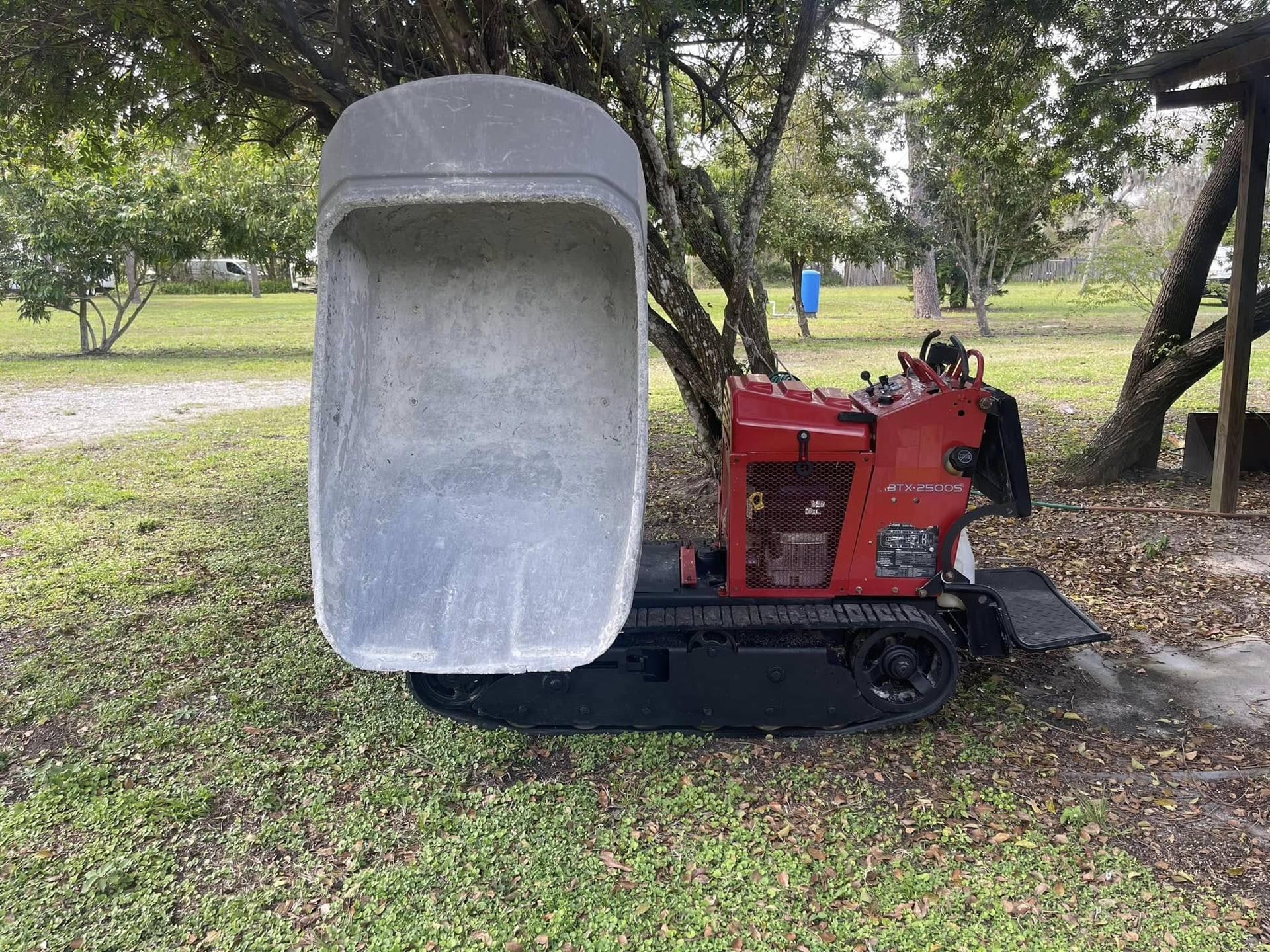Red tracked utility vehicle with a large gray bucket, on a grassy area, trees in the background.
