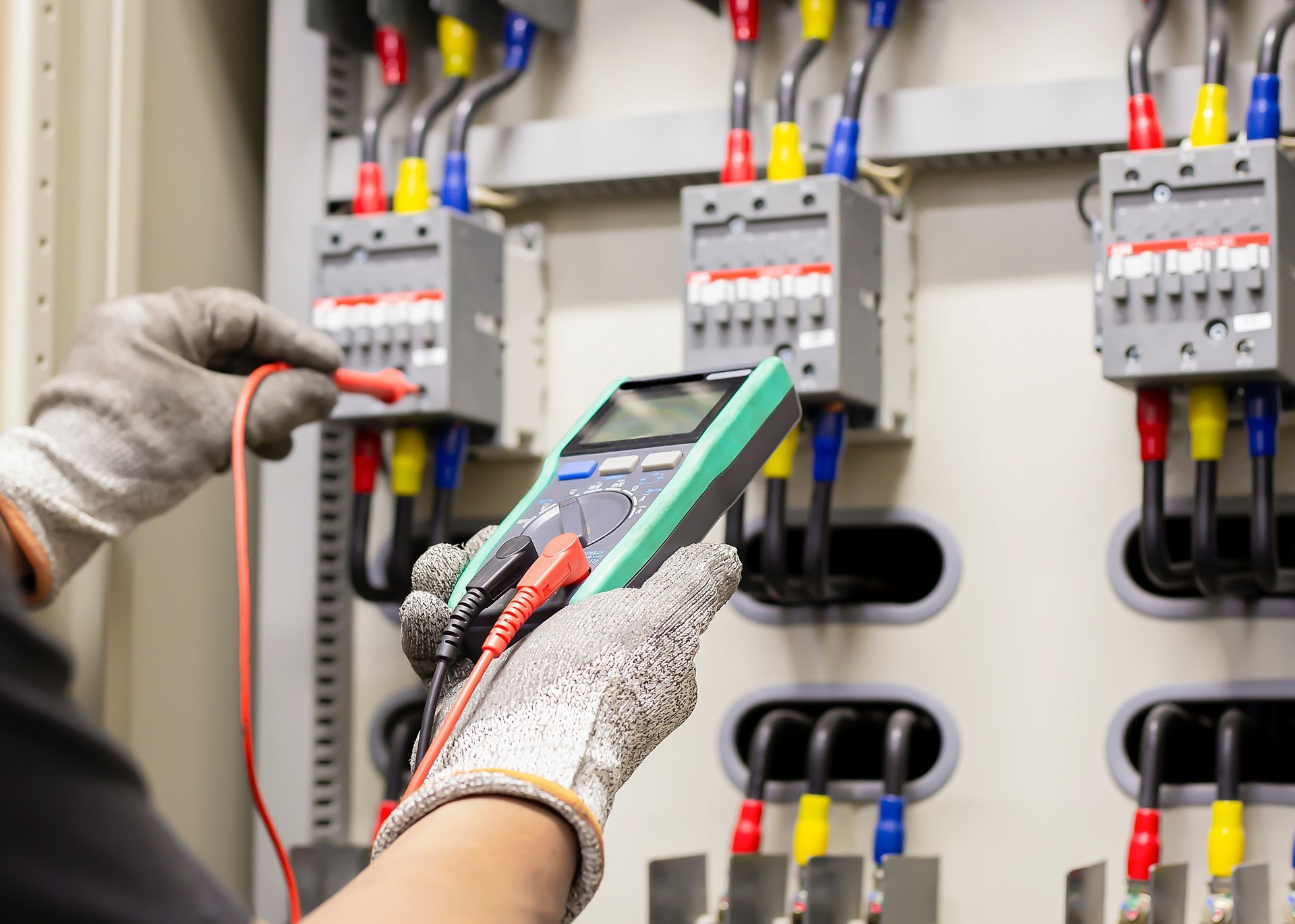 Gloved hands testing electrical wiring in a control panel with circuit breakers and colored cables