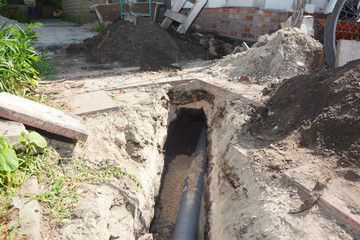 Trench with exposed black pipe under a dirt-covered outdoor worksite, with piles of soil and concrete nearby