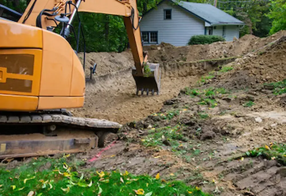 Yellow excavator digging a dirt slope near a house and pile of earth