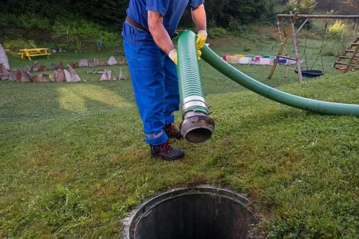 Worker using a green hose to pump water into a ground well in a grassy yard