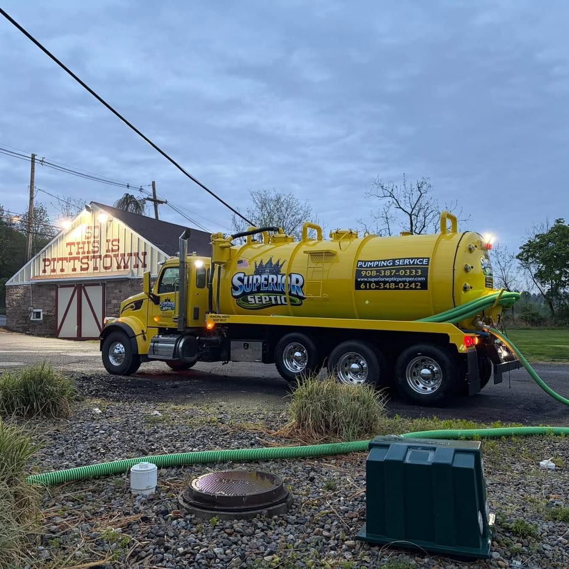 Yellow vacuum truck parked beside a lit building at dusk, with hoses laid across the ground.