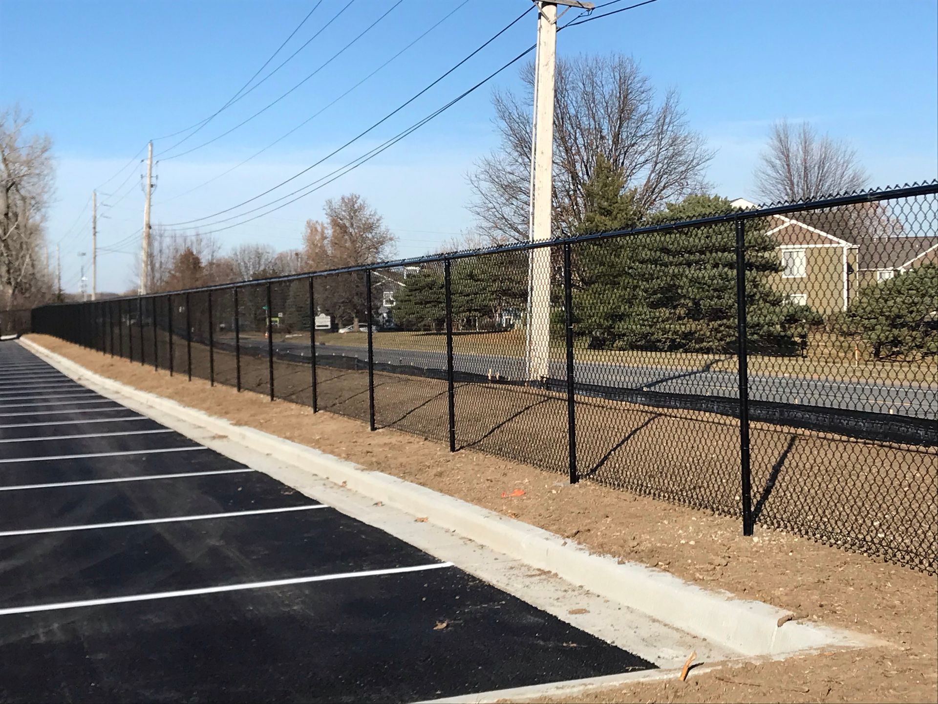 Black chain-link fence bordering a parking lot and yard, under a blue sky with power lines.