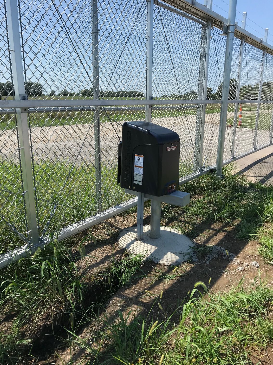 Black gate opener box on a concrete base next to a chain-link fence.