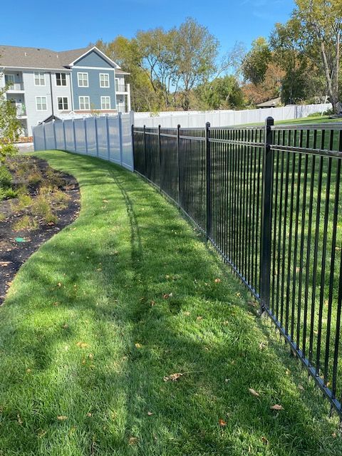 Black metal fence curves along green grass in front of a light blue apartment building on a sunny day.