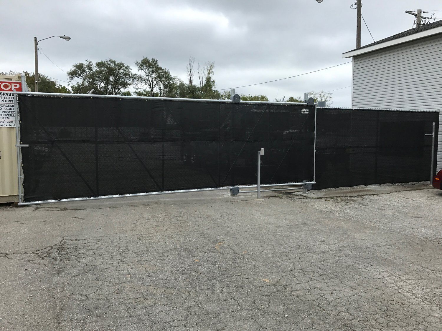 Black privacy fence in front of a building on a gray concrete surface under a cloudy sky.
