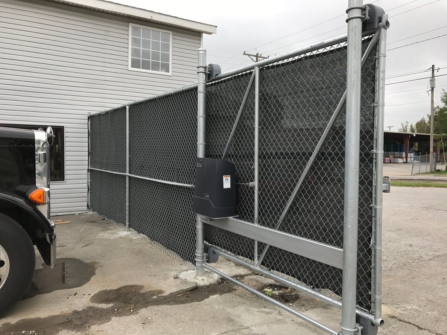 Black security gate, chain-link fence, attached to building; black vehicle parked to the left.