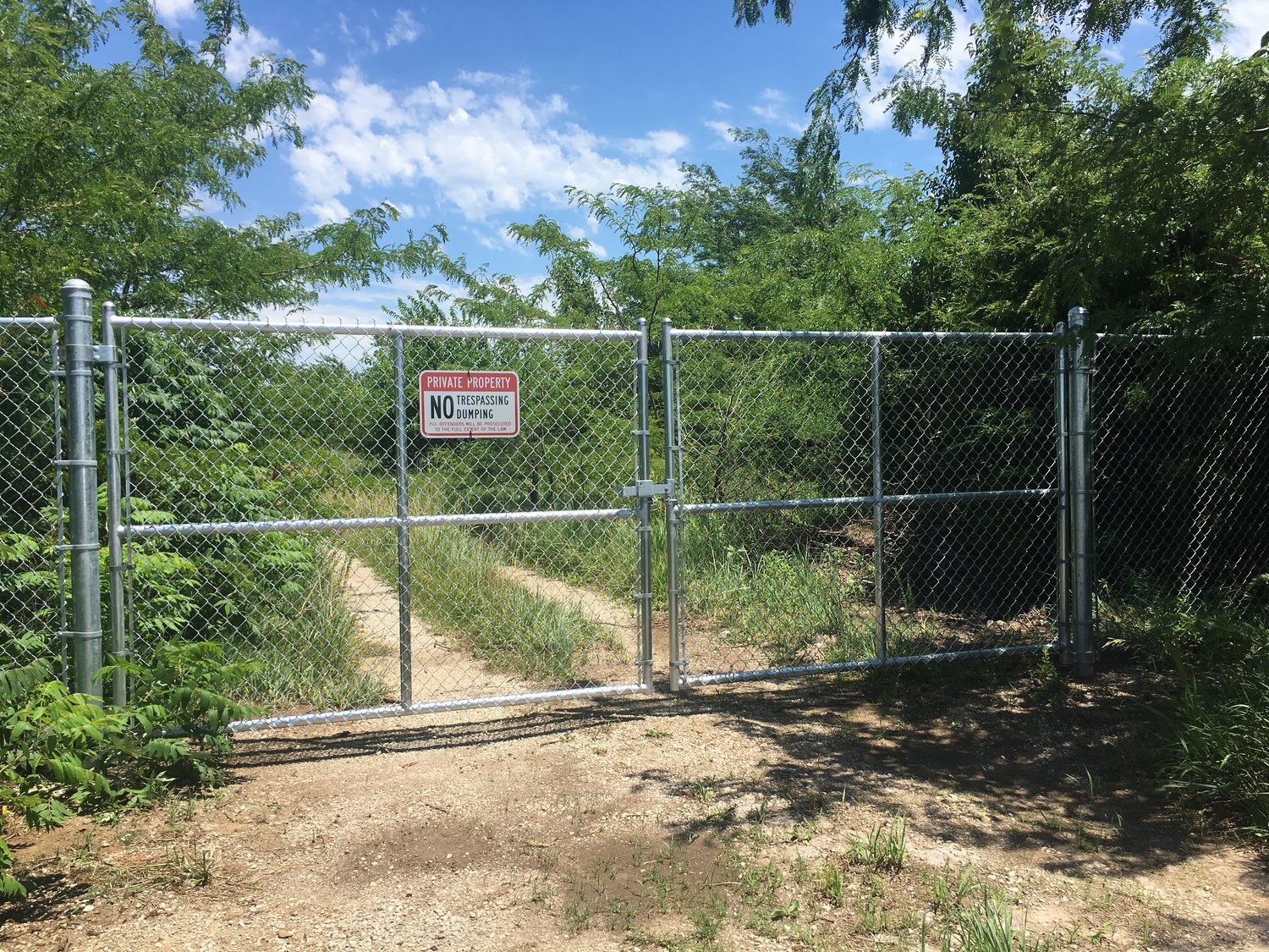 Chain-link fence with a closed gate blocks a dirt path. A warning sign hangs on the gate. Green foliage surrounds it.