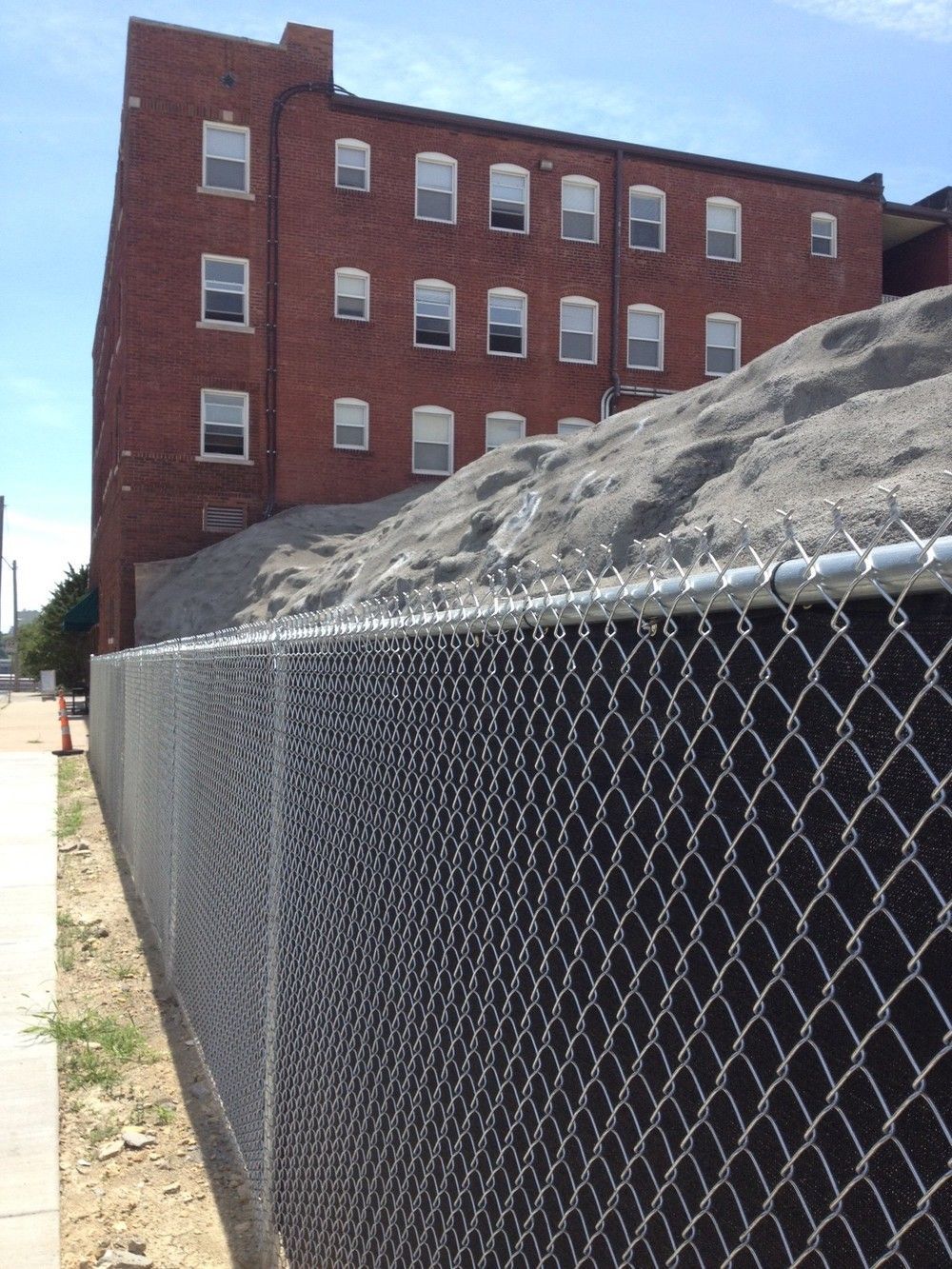 Chain-link fence in front of a pile of gravel next to a brick building with many windows.